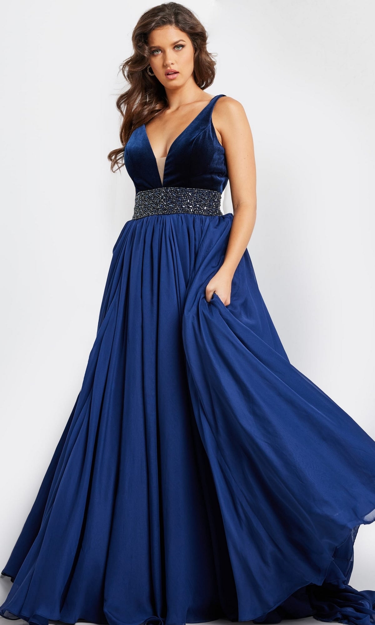 A woman wears the Jovani Belted Long Chiffon Prom Ball Gown 26201, featuring a sleeveless deep blue design with a deep V-neckline, beaded waistband, and a full flowing skirt.