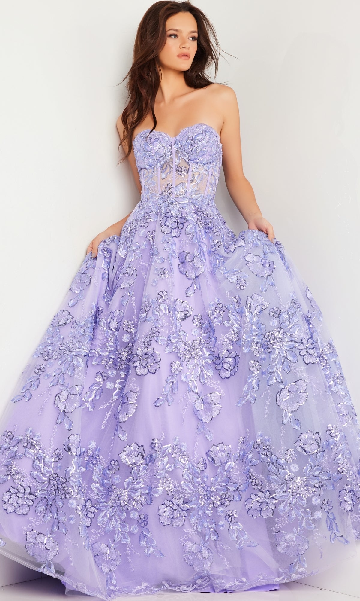A woman models the Jovani Strapless Lilac Long Prom Ball Gown 26223, featuring floral appliqué details, as she stands against a white wall and holds out the sides of her skirt.