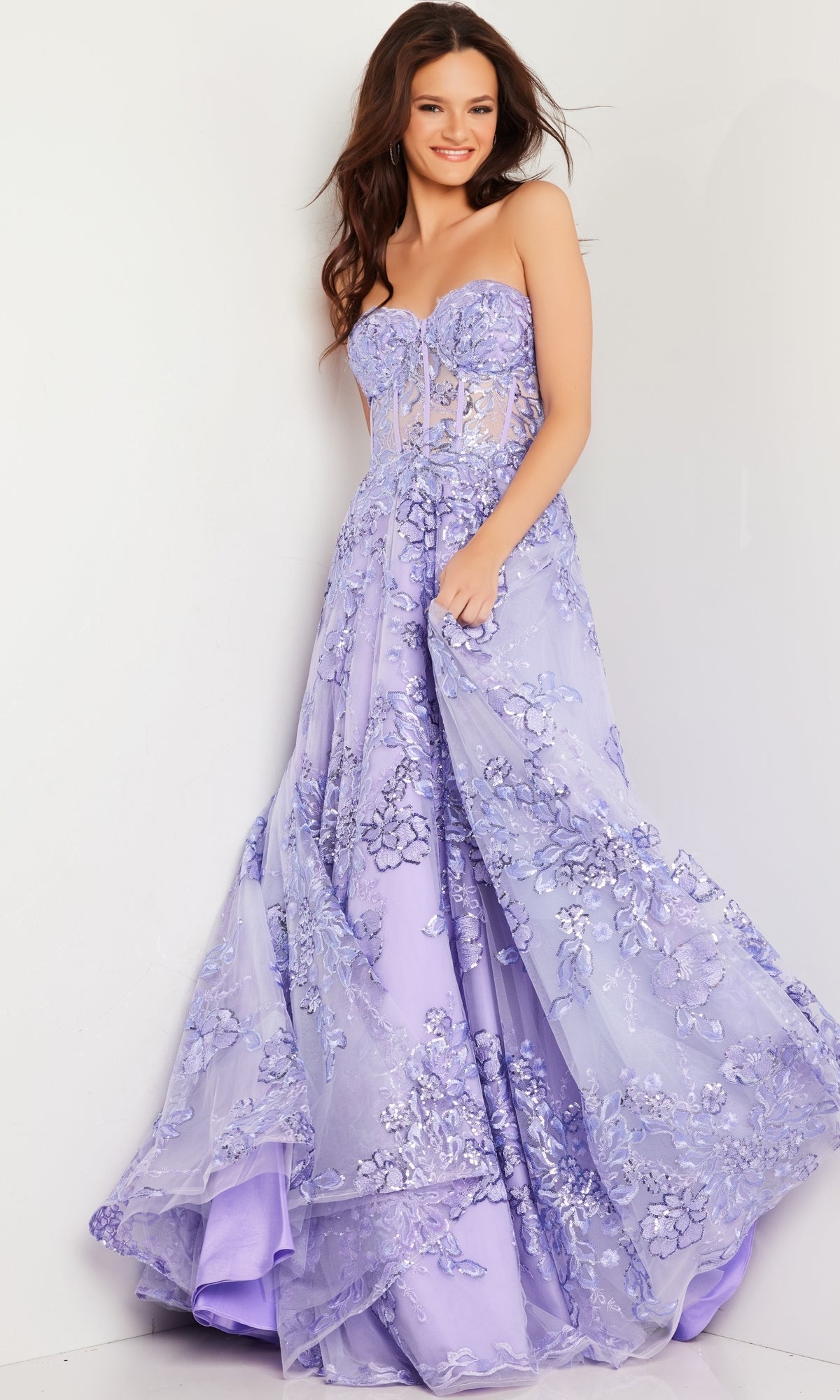 A woman smiles against a white wall, holding her dress while wearing the Jovani Strapless Lilac Long Prom Ball Gown 26223 with delicate floral embroidery.