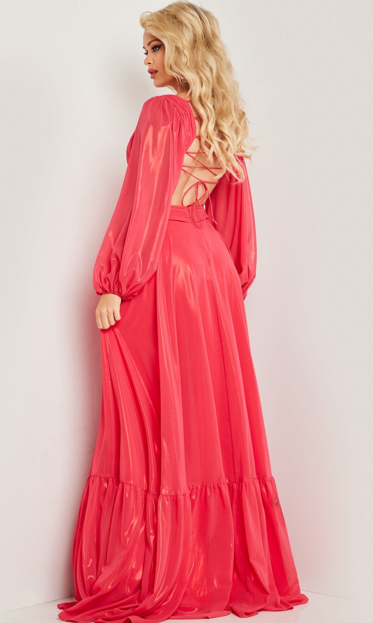 A person with long blonde hair models the Metallic Chiffon Long Coral Formal Dress with Sleeves, which is shiny, floor-length, and features a lace-up back, standing against a plain white wall.