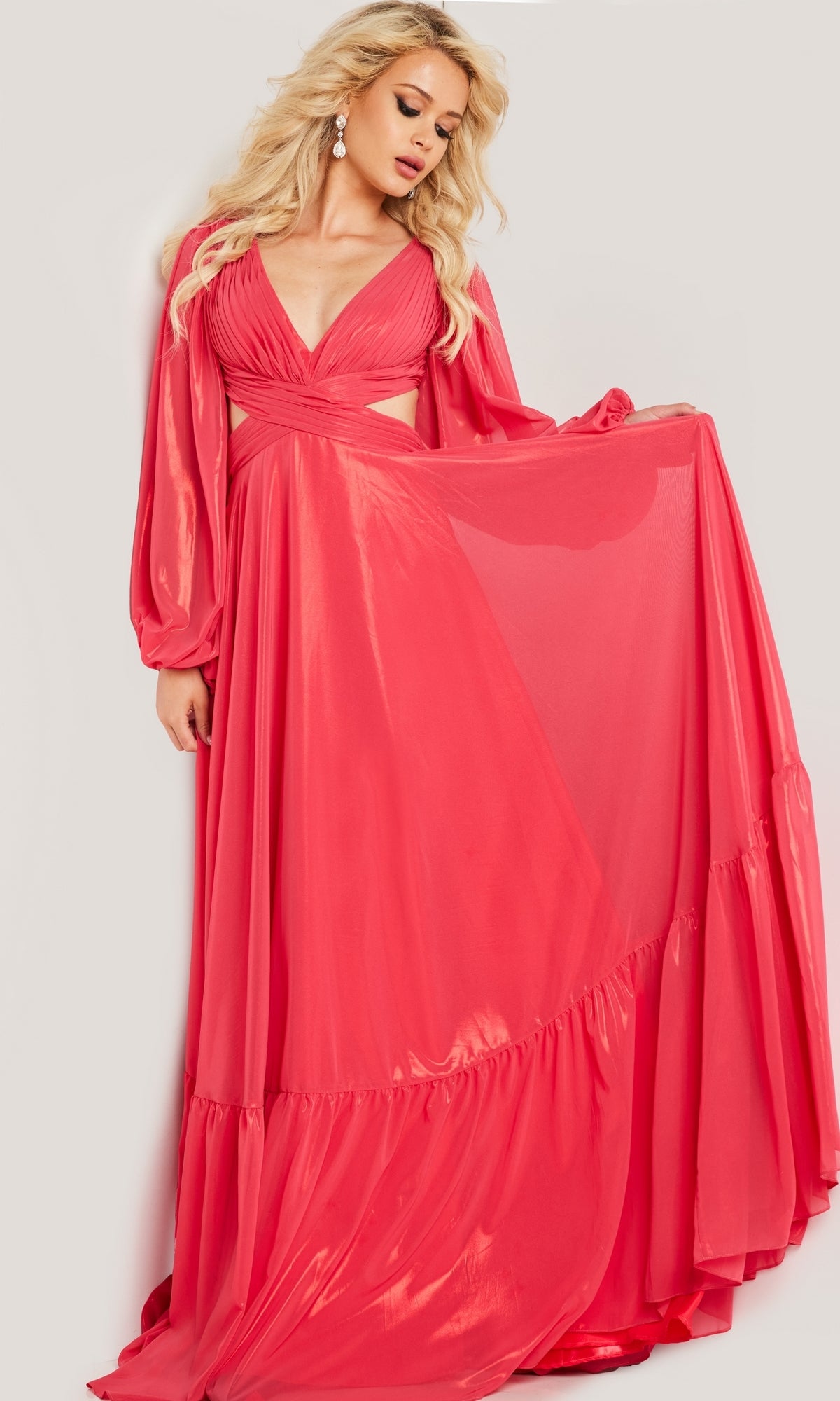 A woman wears the Metallic Chiffon Long Coral Formal Dress with Sleeves by Jovani, featuring shiny fabric, long sleeves, a deep V neckline, side cutouts, and gracefully holds out one side of the skirt.