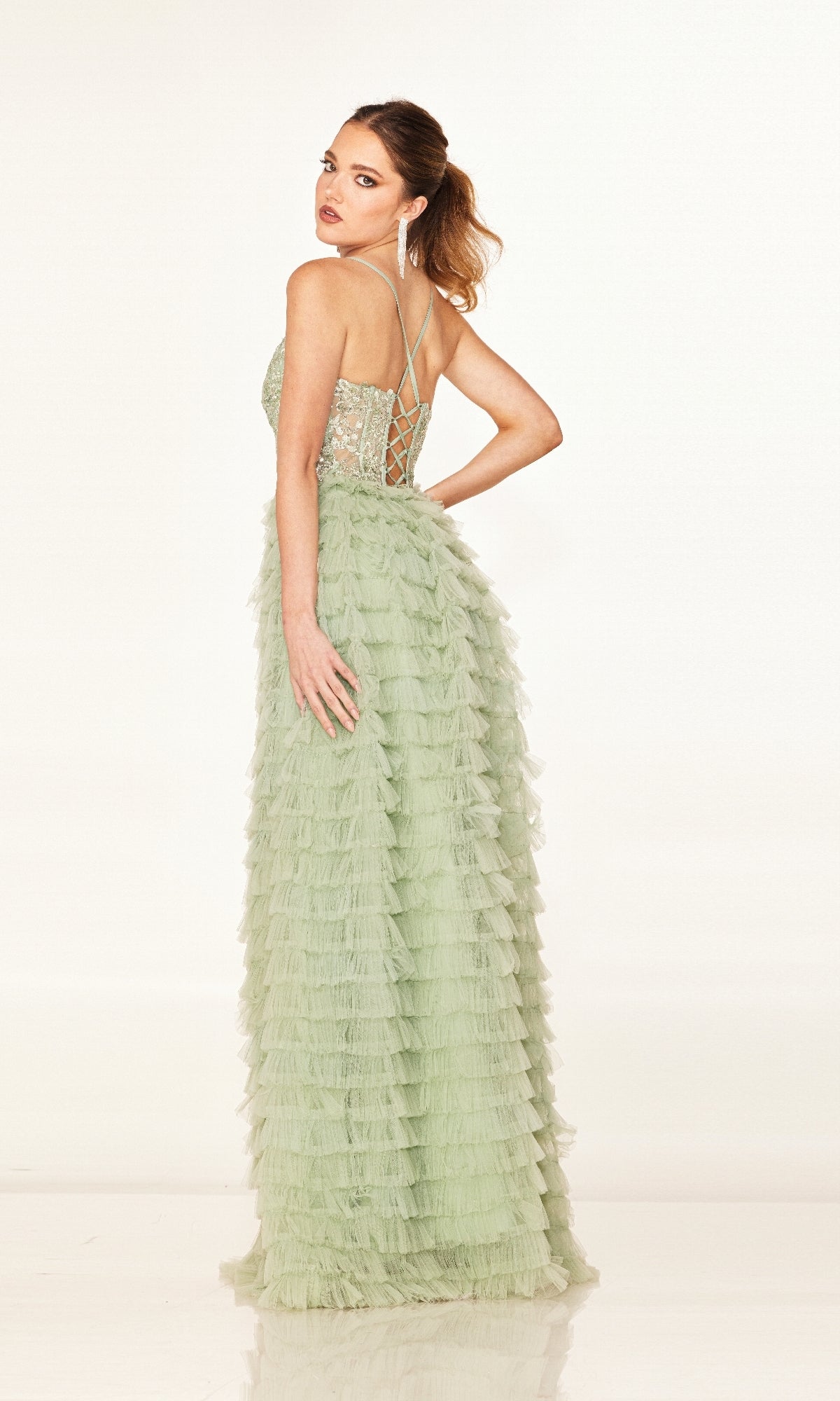 Woman in the Cecilia Couture 2624 long prom dress—light green, A-line with ruffled skirt, sheer corset bodice, and lace-up back—poses with one arm on her hip, looking over her shoulder.
