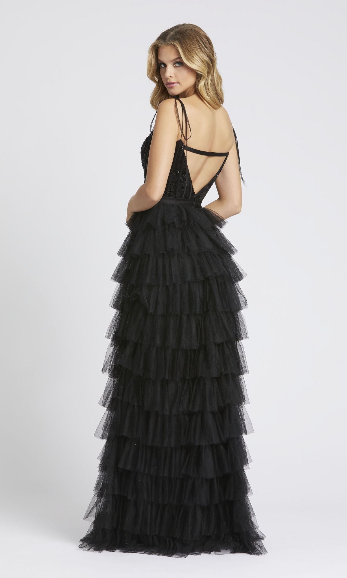 Woman wearing the Long Black Ruffled Prom Dress: Mac Duggal 26281, which features a sleeveless design, crisscross straps, and a tiered ruffled skirt, standing and looking over her shoulder against a plain backdrop.