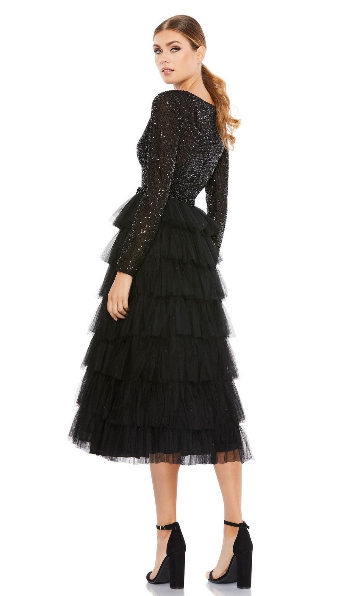 A woman stands in profile, head turned toward the camera, wearing the Mac Duggal 26299 knee-length wedding guest dress with a black sequined top and tiered tulle skirt, against a plain white background.
