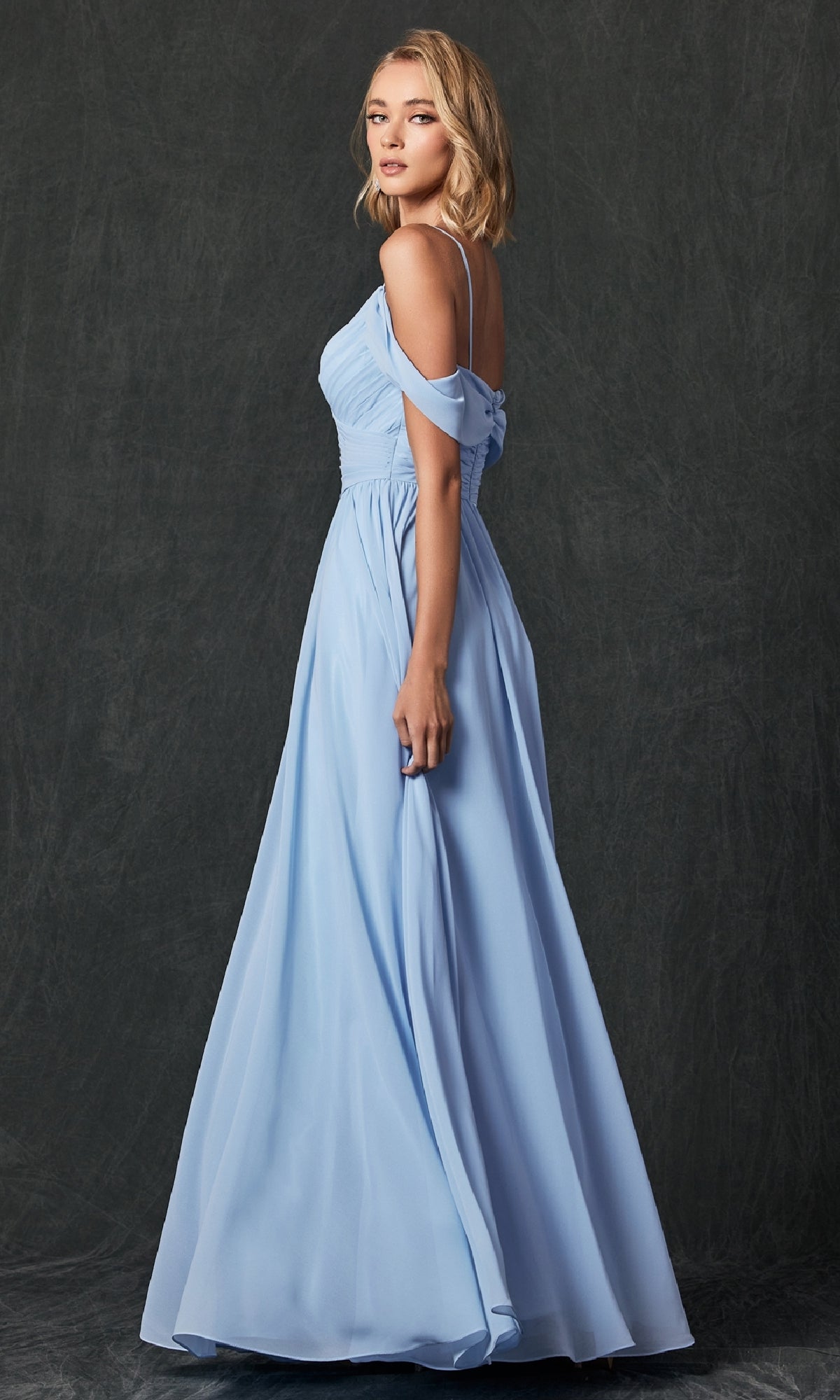 A woman stands in profile against a dark, textured background wearing the Juliet 262 Long Prom Dress, a floor-length light blue A-line gown with off-the-shoulder straps.