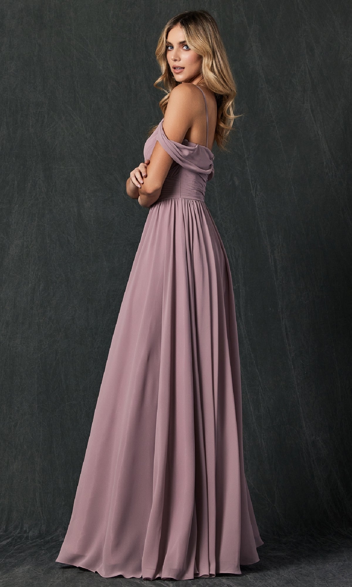 A woman wearing the Juliet 262 long prom dress in mauve, featuring an off-the-shoulder a-line design, stands sideways against a dark, textured background.