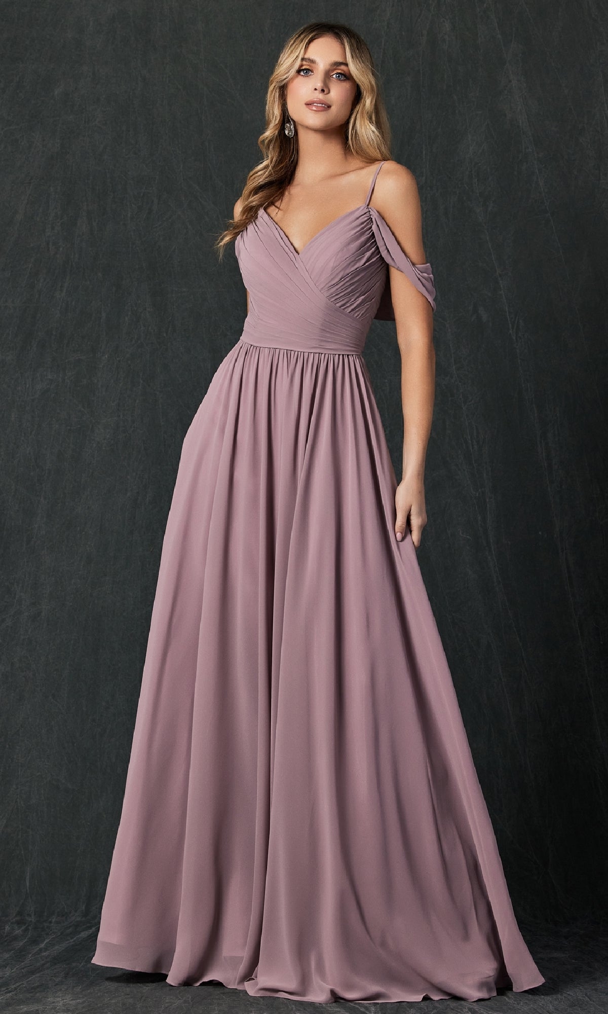 A woman models the Juliet 262 long prom dress, a mauve, floor-length A-line gown with off-the-shoulder straps, pleated bodice, and flowing skirt, posed against a dark textured background.