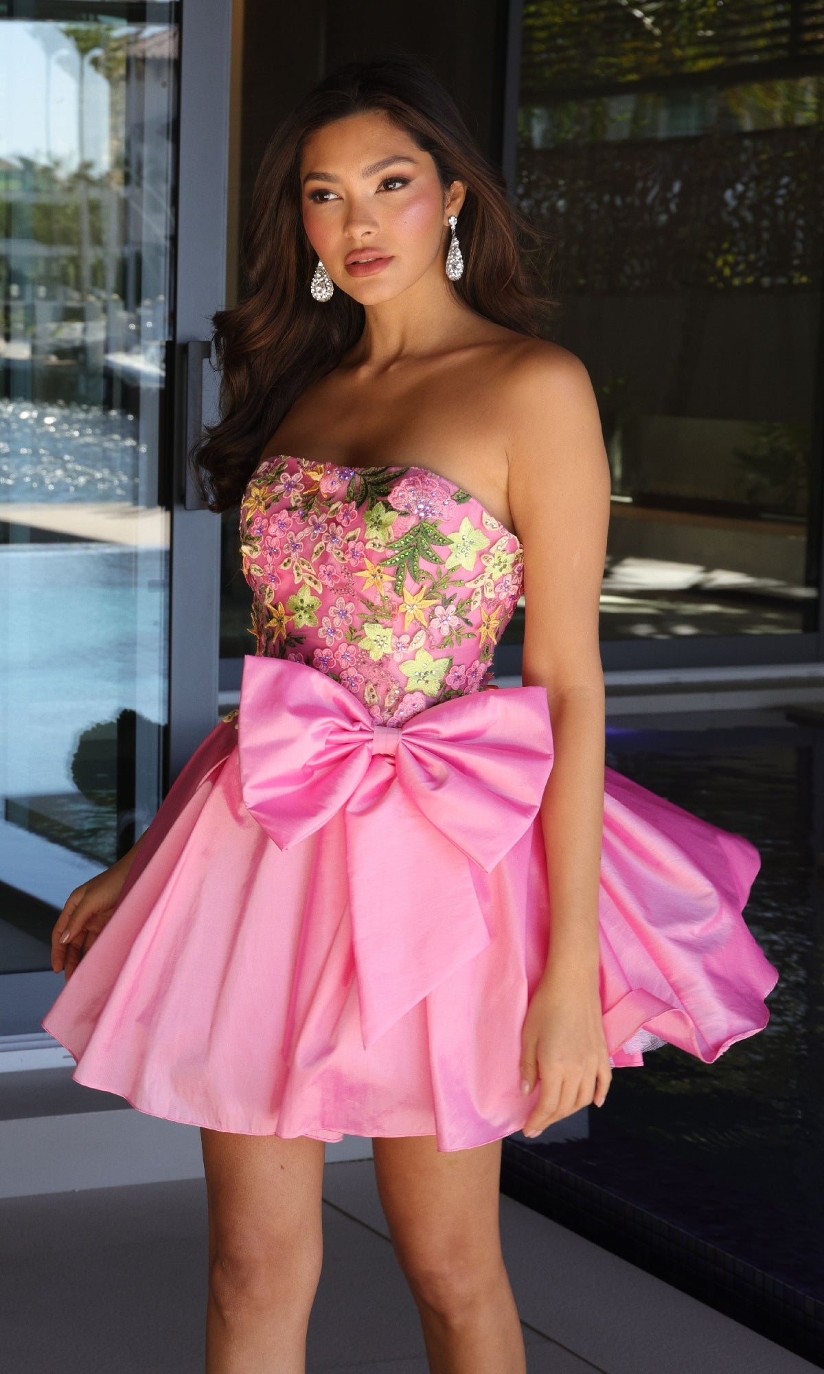 A woman stands indoors wearing the Ava Presley 26347 short homecoming dress, featuring a strapless floral-embroidered bodice and large bow, paired with drop earrings.