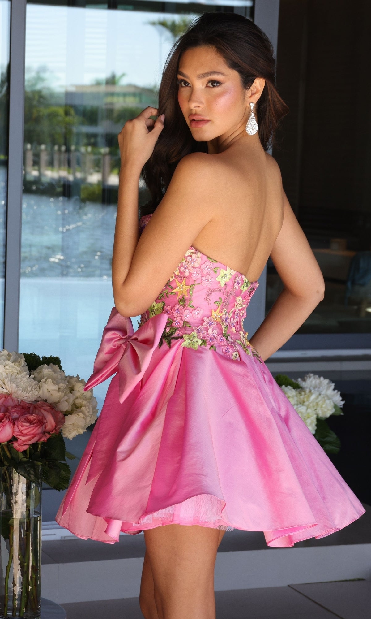A woman stands indoors in the Ava Presley 26347 short homecoming dress, featuring a strapless floral-embroidered bodice and a large bow. She looks over her shoulder with flowers and a window view in the background.