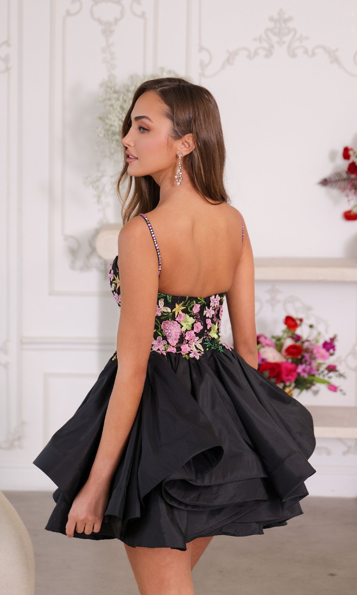 A woman indoors looks over her shoulder wearing the Ava Presley 26348 short homecoming dress, featuring a sleeveless black design with a layered skirt and floral-embroidered bodice. Flowers can be seen in the background.