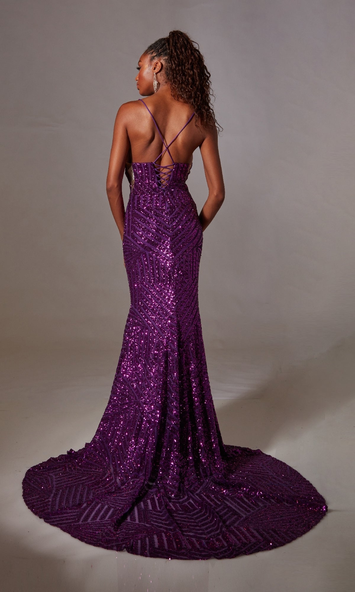 A woman wearing the Aerie Couture 263 Lace-Up Long Sequin Prom Dress, a floor-length purple gown with a train and criss-cross back straps, stands against a neutral background.