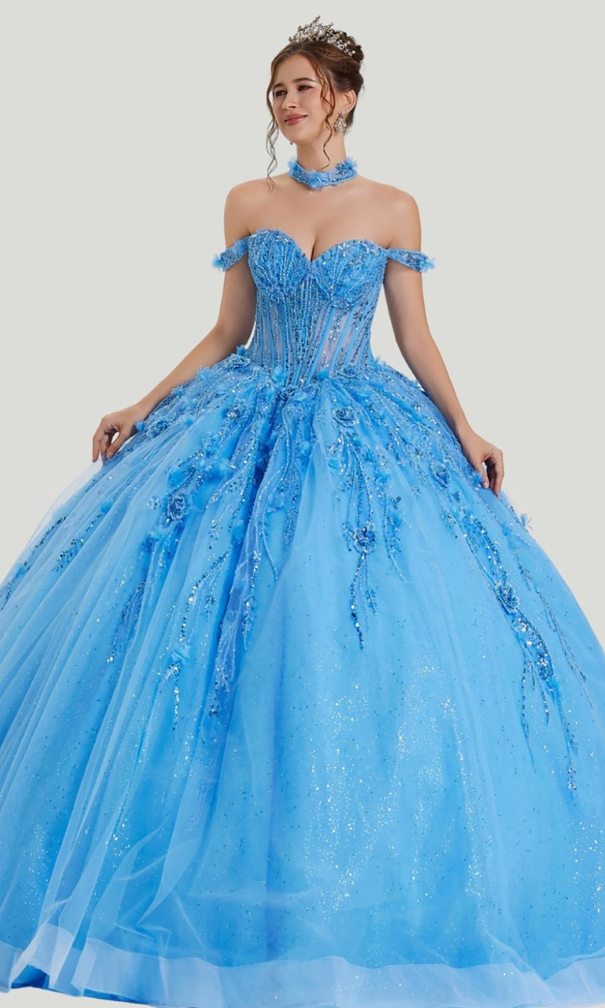 A woman wearing the Quinceanera Dress 26416 by Stella Couture in Azure, featuring an off-shoulder, embellished corset bodice and a glitter tulle skirt adorned with delicate floral details, stands against a plain background.
