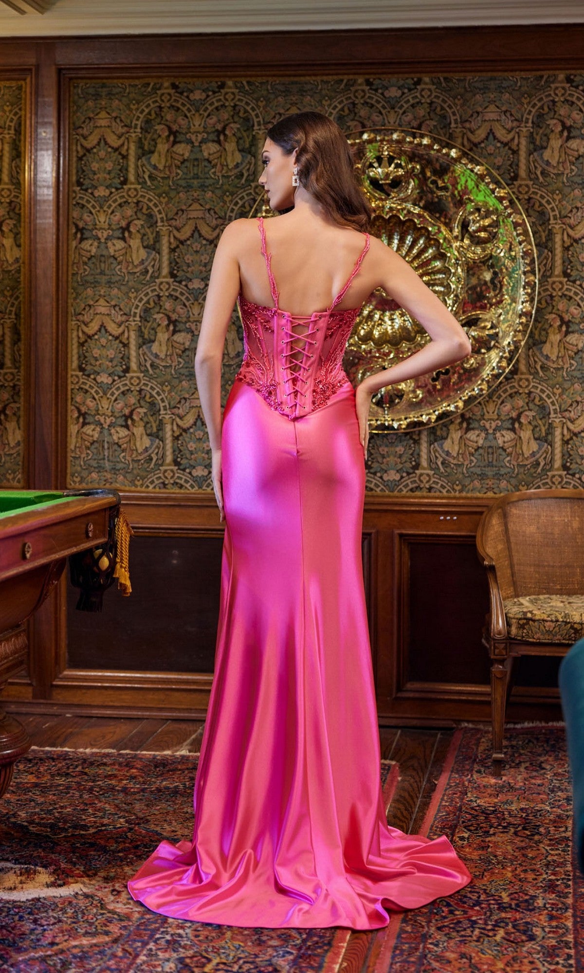 A woman faces away from the camera in a wood-paneled room with ornate decor, wearing the Juliet JT2645K Long Prom Dress—a fitted, floor-length pink satin gown.