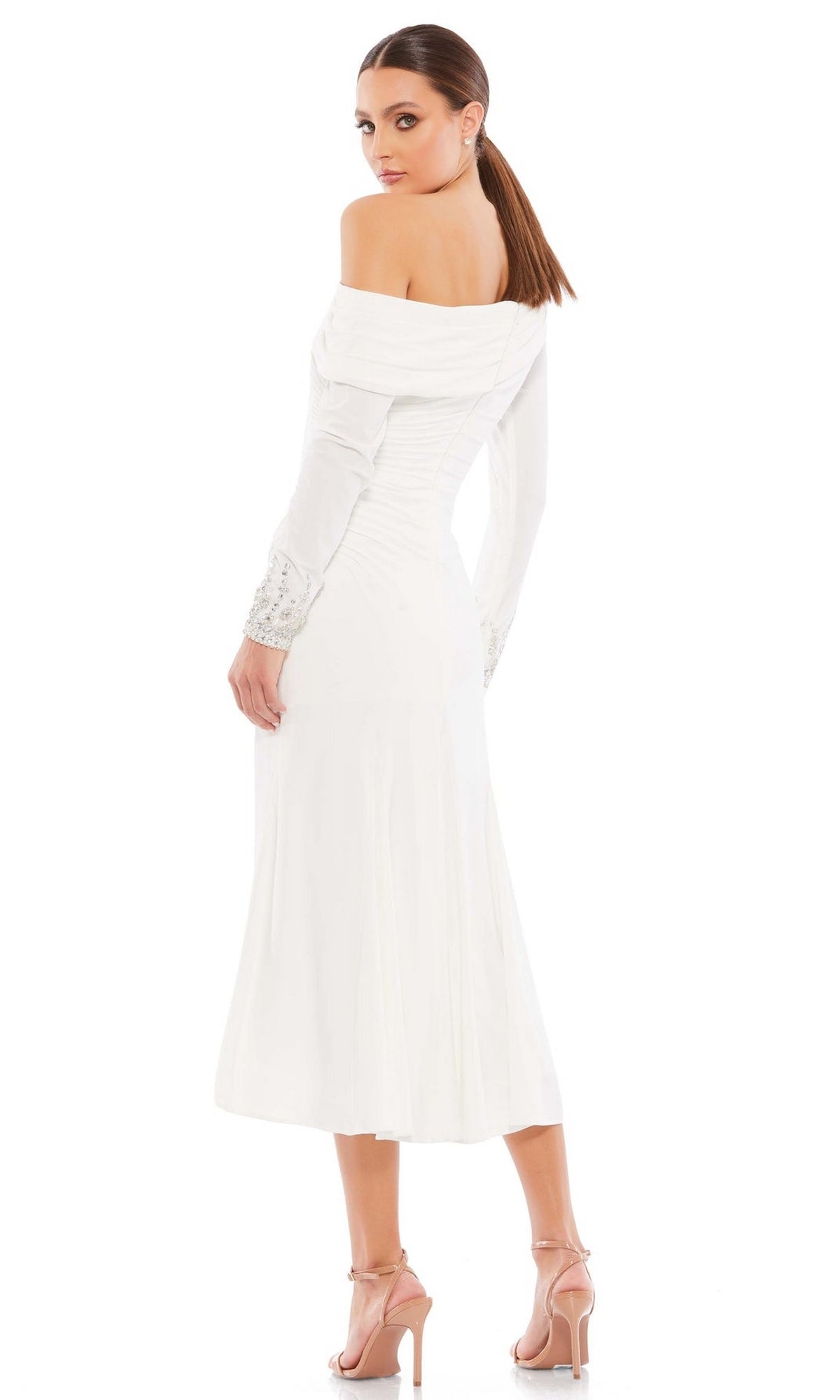 A woman models the Knee Length Party Dress 26485 by Mac Duggal, a drop shoulder white midi dress with embellished cuffs and long sleeves, paired with nude high-heeled sandals, standing against a plain white background.