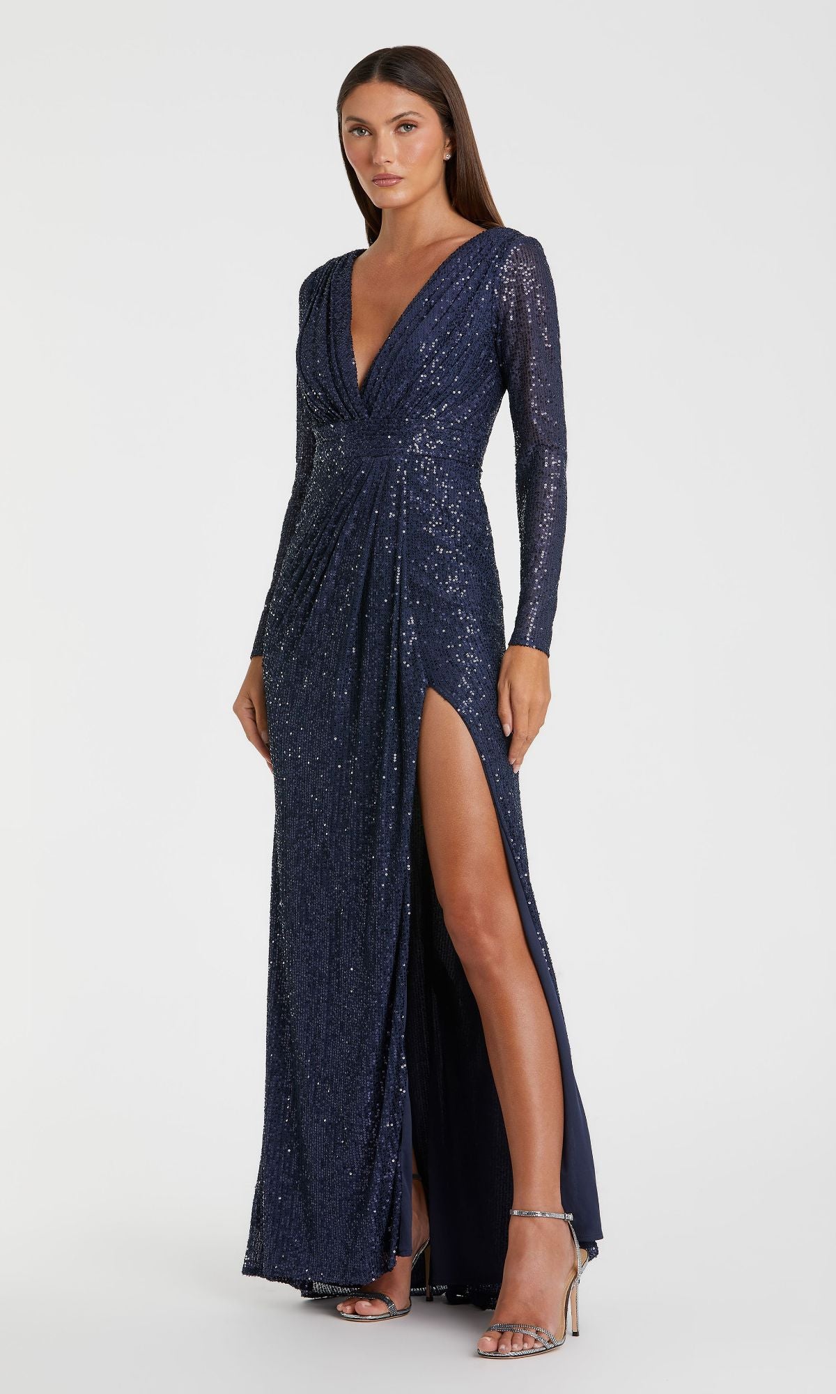A woman in the Mac Duggal 26490 long-sleeve formal dress with navy blue sequins, a deep V-neckline, and a high slit, paired with silver high-heeled sandals.