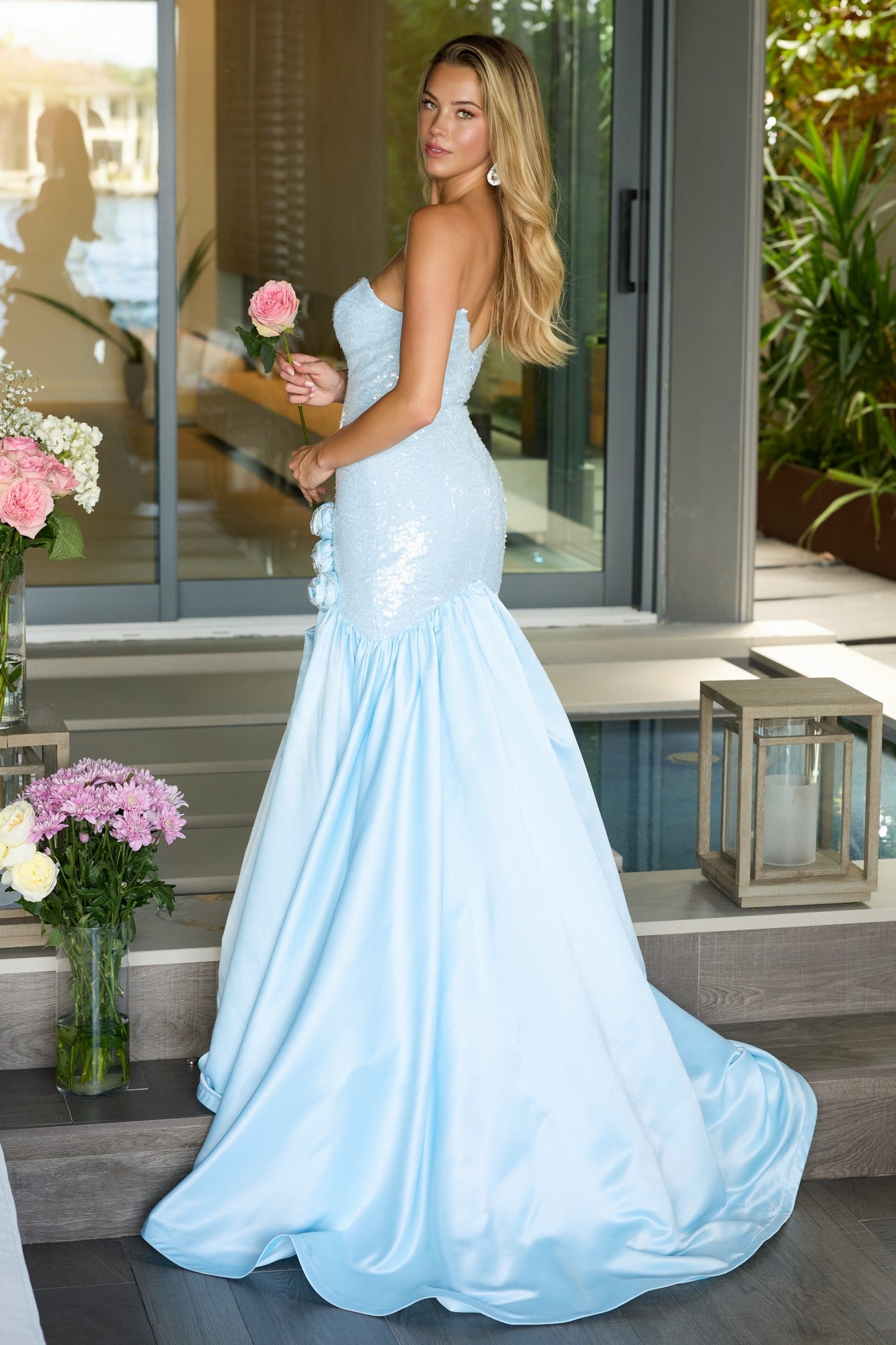 A woman in the Ava Presley 26520 long prom dress, featuring a strapless mermaid silhouette and sequin fitted bodice, holds a pink rose while standing indoors near glass doors and floral arrangements.