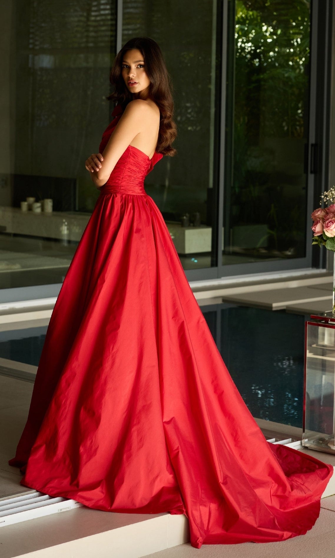 A woman stands indoors near a pool, looking over her shoulder in the strapless, floor-length red Ava Presley 26524 Long Prom Dress.