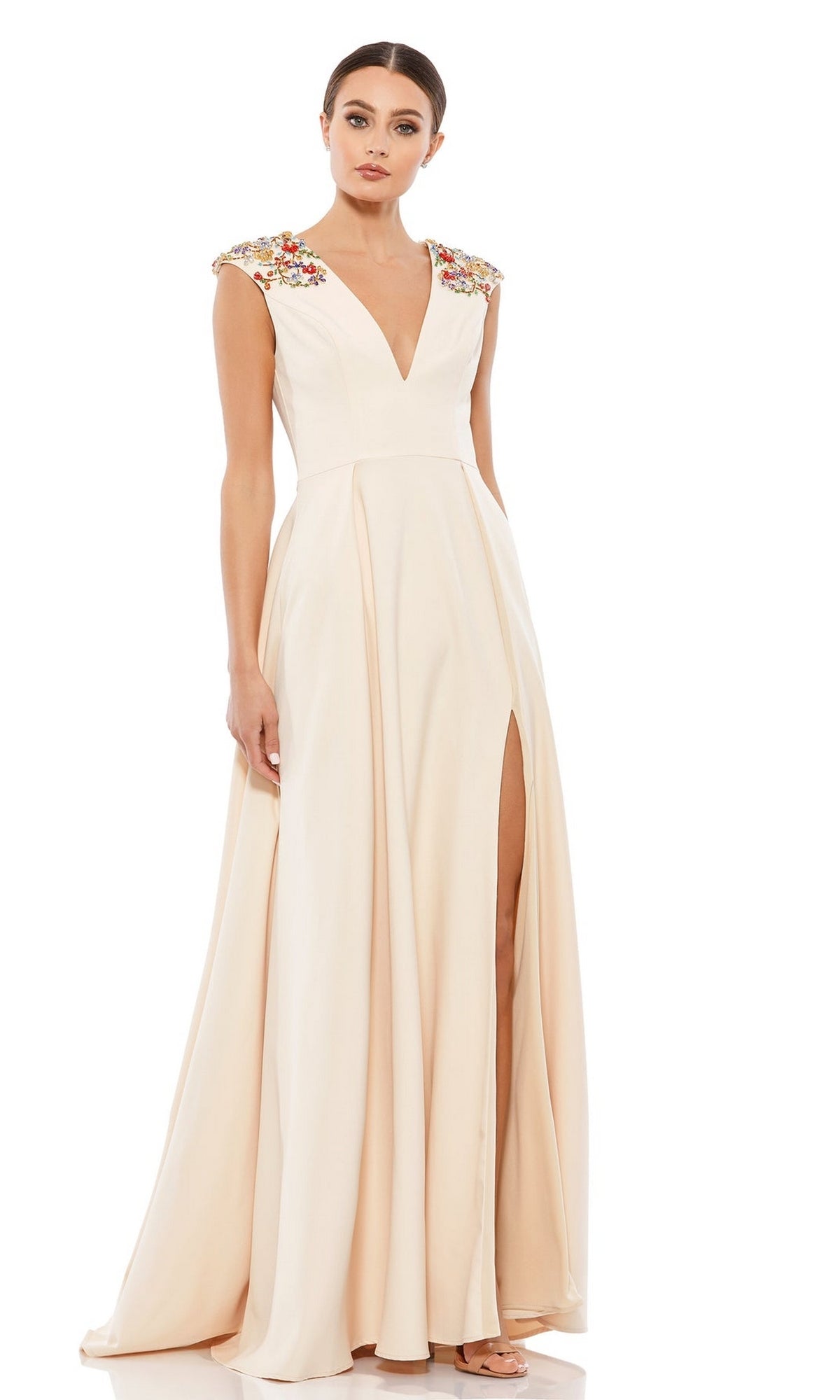 A woman models the Mac Duggal 26540 Cap-Sleeve Long Formal Dress with a floor-length cream skirt, high slit, deep v-neck, embellished shoulders, and stands against a white background.