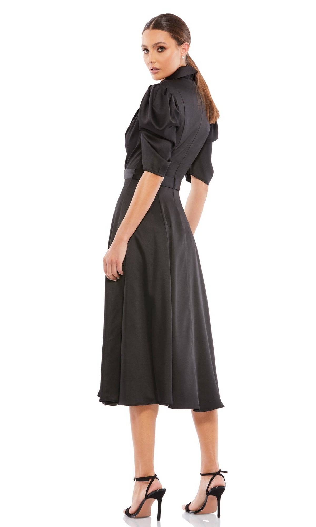 Woman wearing the Mac Duggal 2628 Short Sleeve Wedding Guest Dress— a black, belted, knee-length midi dress with puffed sleeves— and black high-heeled sandals, standing and looking over her shoulder.