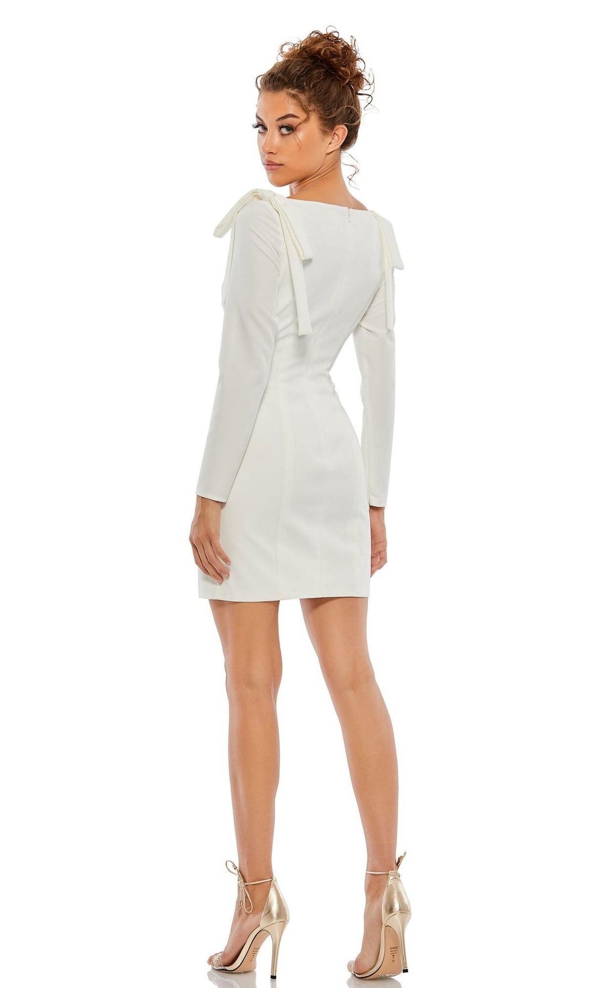 A woman stands facing backward in the Long-Sleeve Short White Semi-Formal Dress 26643, featuring bow shoulder details and paired with metallic high heels, reminiscent of Mac Duggal style, against a white background.