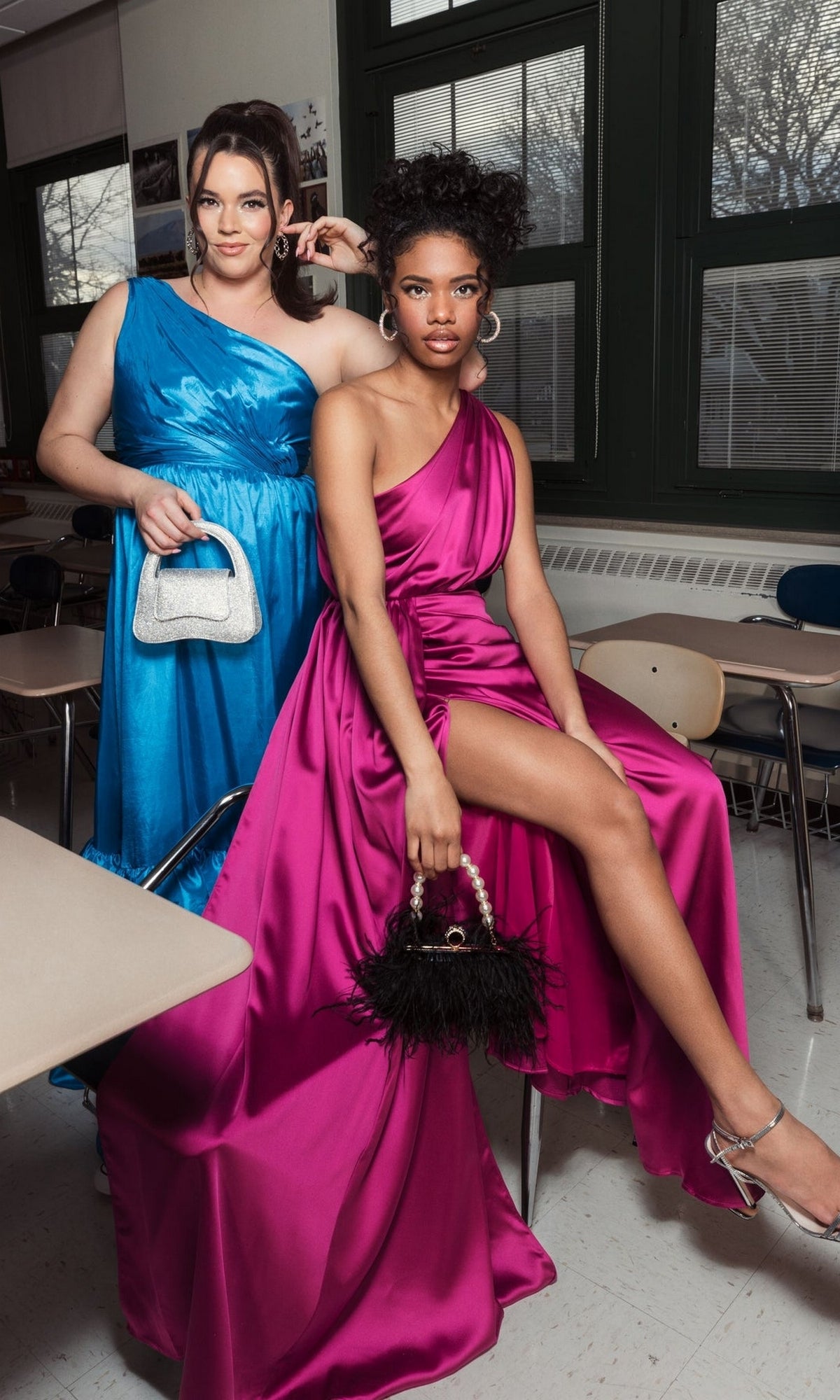 Two women in formal gowns pose with handbags among classroom desks—one wears a blue Mac Duggal 26654 One-Shoulder Long Formal Dress, while the other is in magenta.