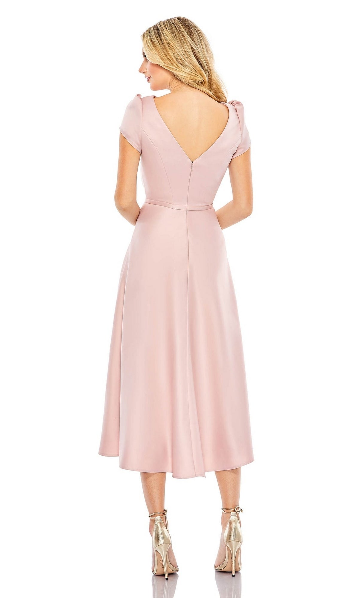 A woman with blonde hair stands facing away, wearing the Short Wedding Guest Dress 26663 by Mac Duggal—a blush pink, A-line knee-length dress with short sleeves and a V-shaped back—paired with metallic high-heeled sandals.