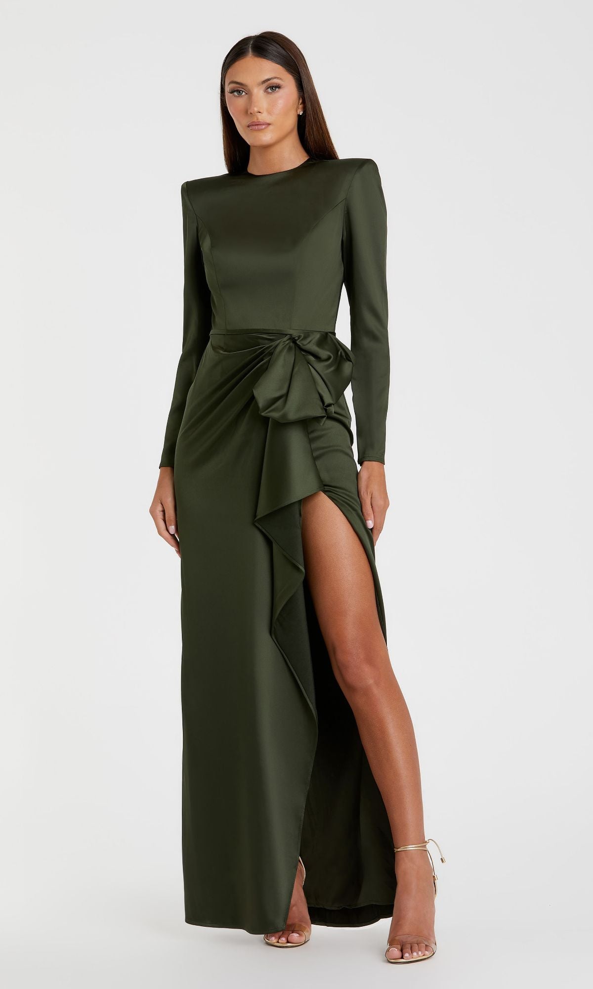 Woman models the Mac Duggal 26676 Bold-Shoulder Long Formal Dress in dark green, with bra-friendly long sleeves, a waist bow detail, and high slit, set against a plain white background.