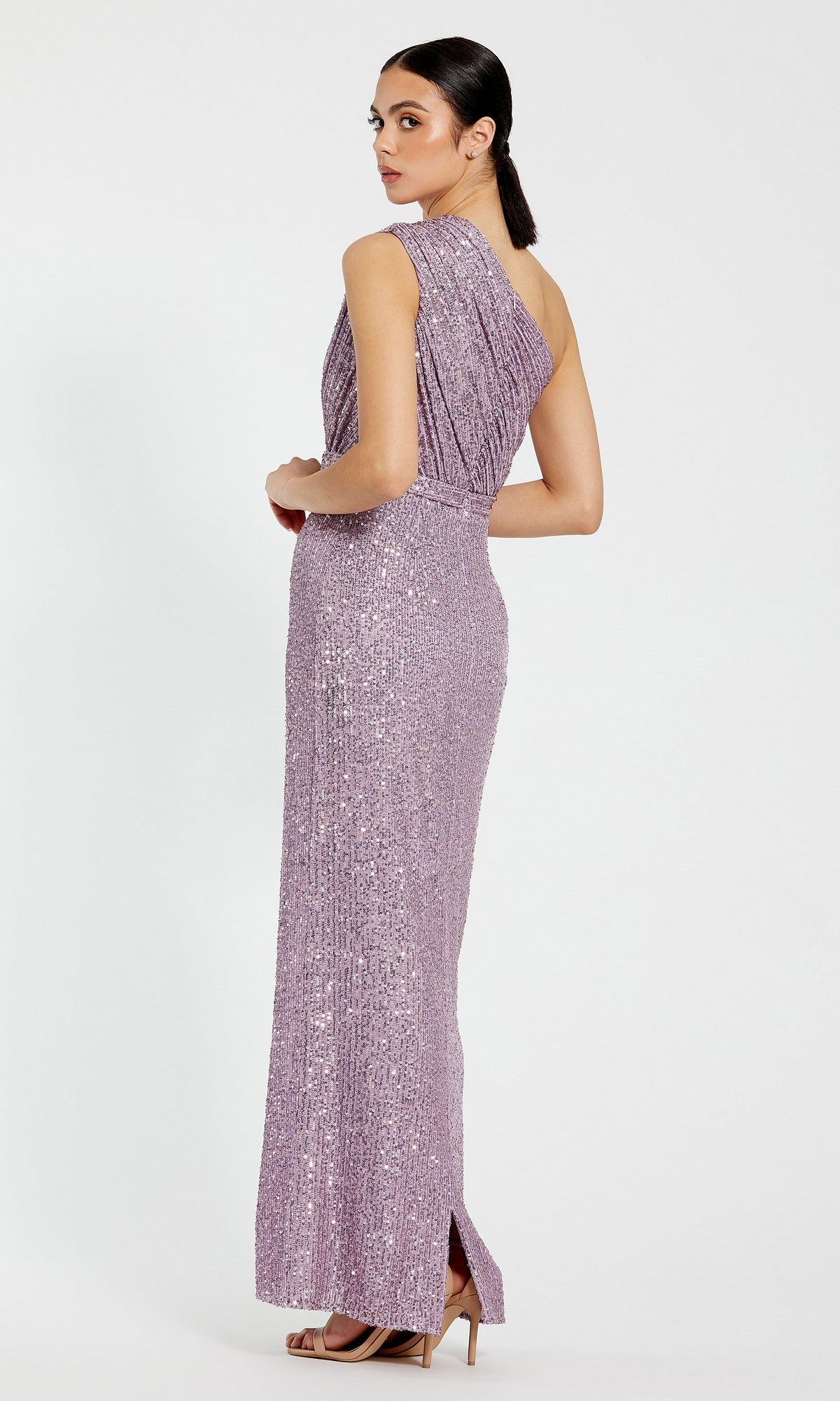 A woman stands in a studio wearing the Mac Duggal 26694 one-shoulder floor-length sequin formal dress in lavender, paired with beige heels, looking over her shoulder.