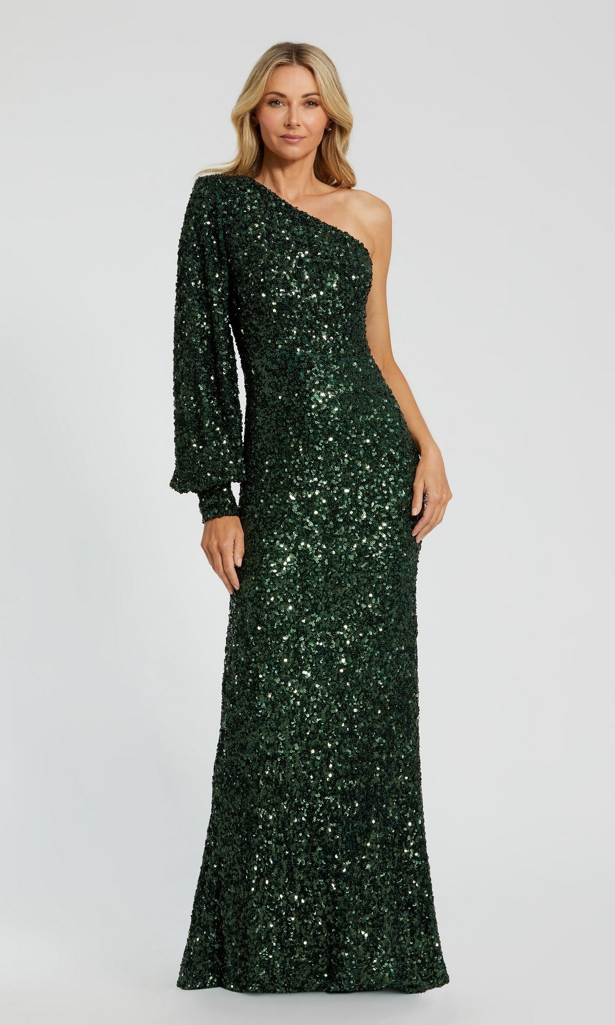 An elegantly dressed woman poses against a plain, light-gray background wearing the Mac Duggal 26717 One-Sleeve Sequin Formal Gown in emerald, featuring a stunning one-shoulder design with a single long sleeve.