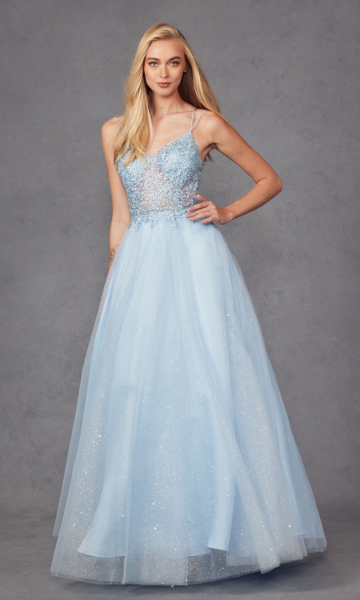 A woman stands against a gray background wearing the Glitter-Tulle Long Corset-Back Prom Ball Gown 268: a light blue, floor-length dress featuring a sheer, beaded bodice with sparkling details.