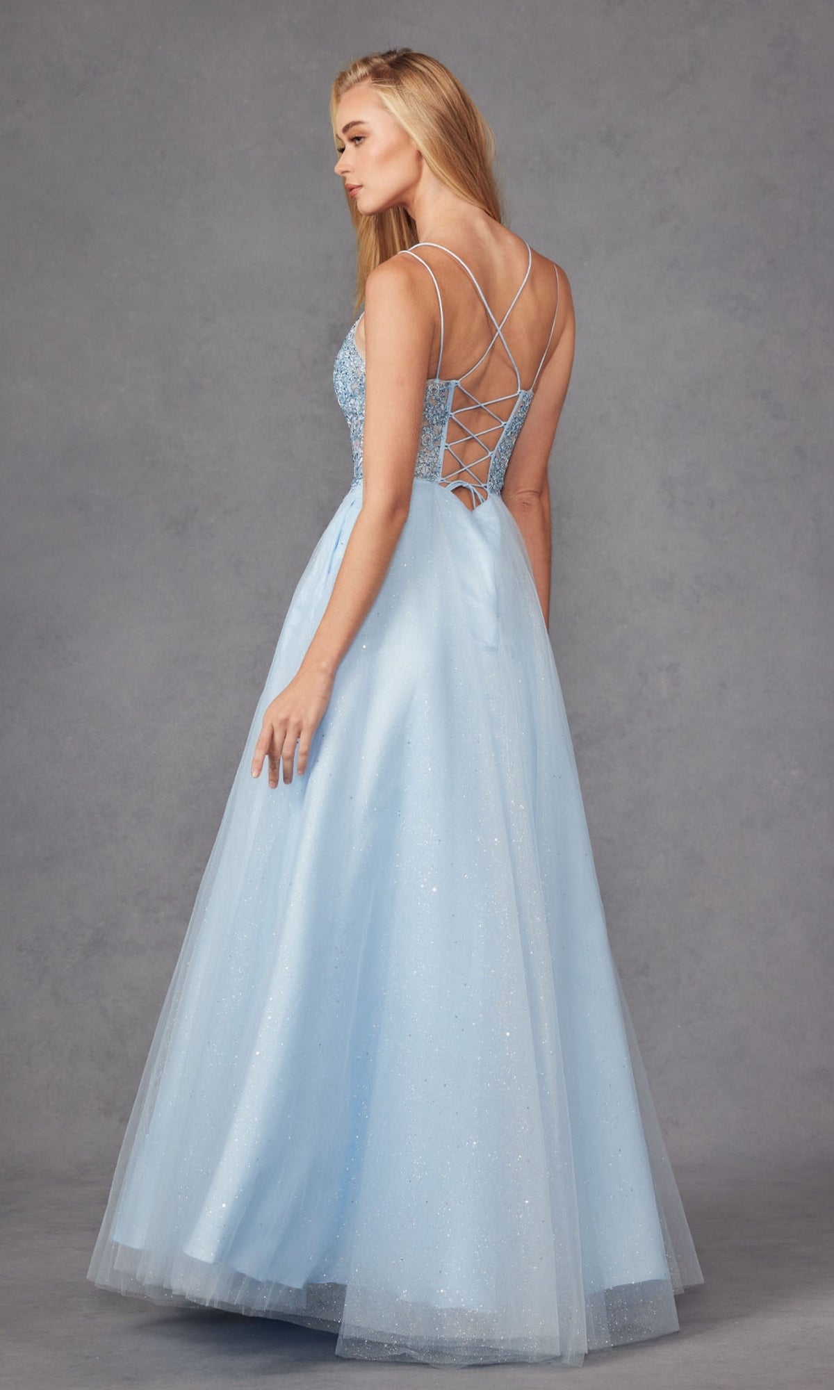 A woman wears the Glitter-Tulle Long Corset-Back Prom Ball Gown 268, an affordable, light blue, floor-length dress with a lace-up back and thin crisscross straps, standing against a gray background.