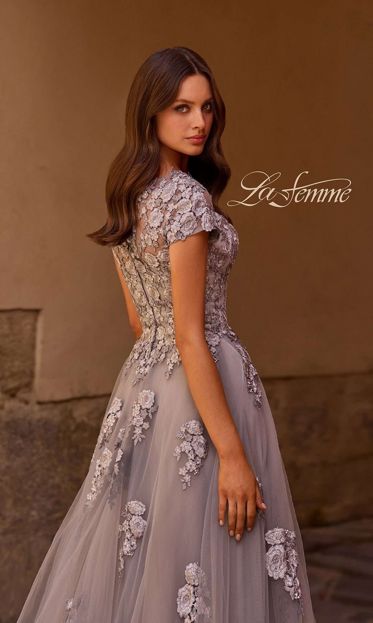 A woman stands sideways, looking over her shoulder against a neutral background, wearing the La Femme 26907 long formal dress—a sheer gown adorned with floral embroidery and embellished lace appliques. The “La Femme” logo appears near her head.