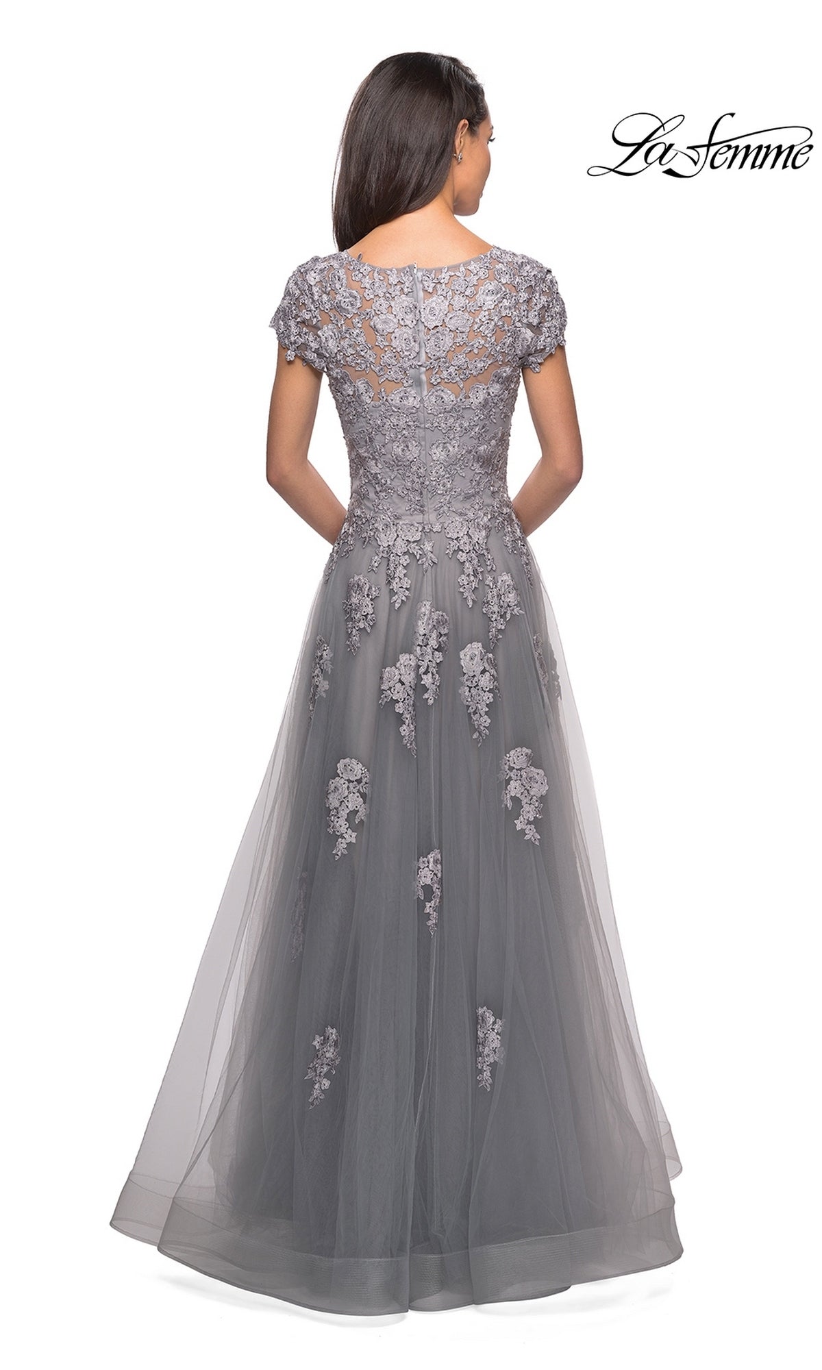 A woman faces away, dressed in the La Femme 26907 long formal gown—a floor-length gray lace dress featuring an elegant A-line silhouette and short sleeves adorned with floral embroidery. The upper bodice and back are embellished with delicate lace appliqués, while the lightly decorated tulle skirt adds a refined finishing touch.