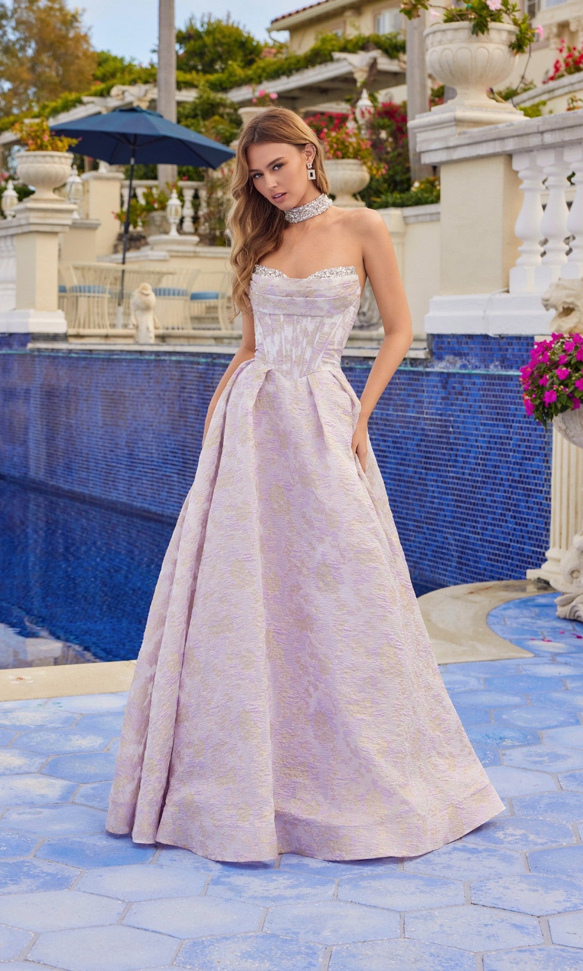 A woman wearing the Long Prom Dress: Juliet JT2693A, a strapless jacquard A-line gown with a matching choker, stands by a blue-tiled pool, flowers and a building in the background.