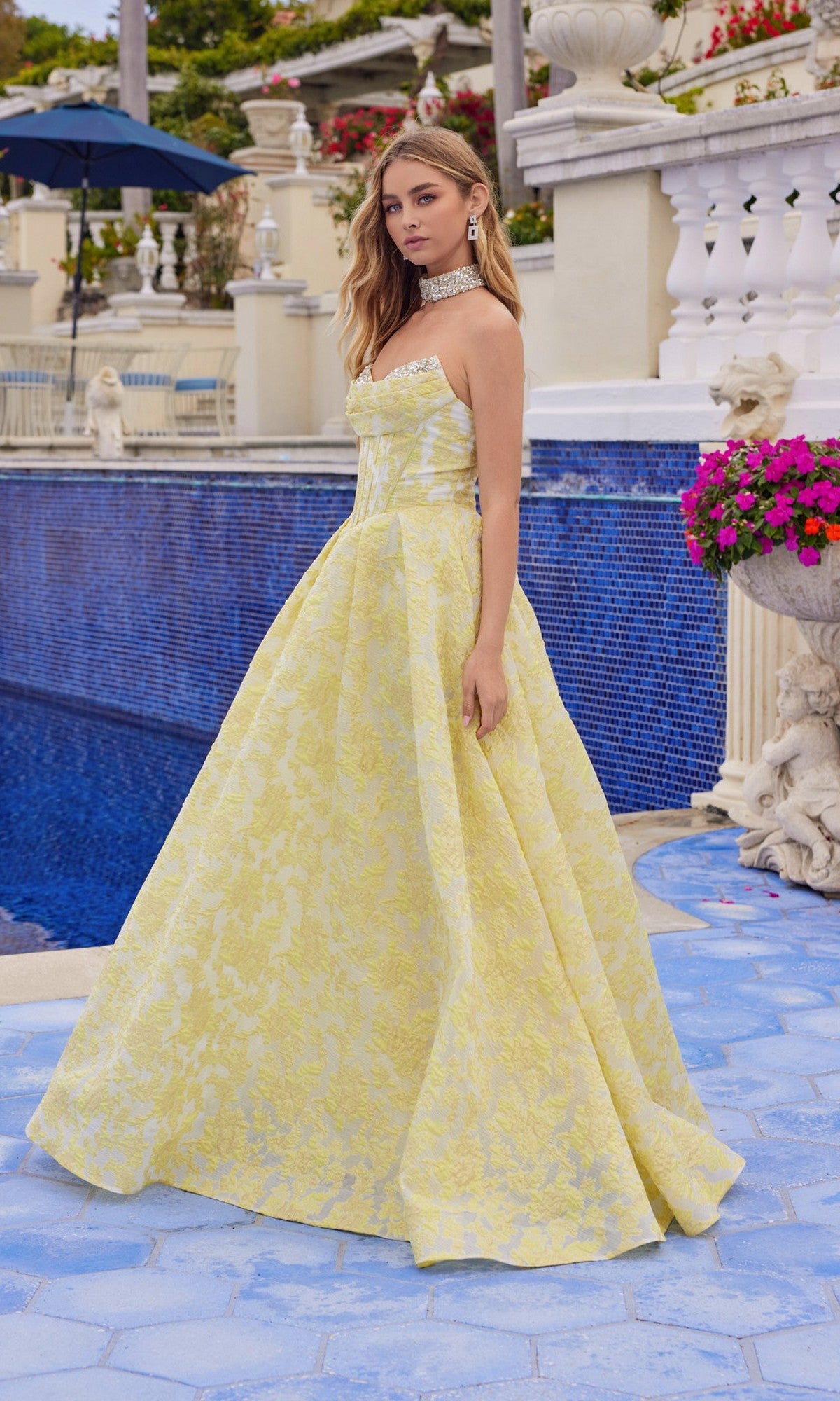 Wearing the Juliet JT2693A long prom dress with a strapless jacquard corset bodice and full A-line skirt, a woman stands by a blue-tiled pool framed by white railings, pink flowers, and lush greenery.