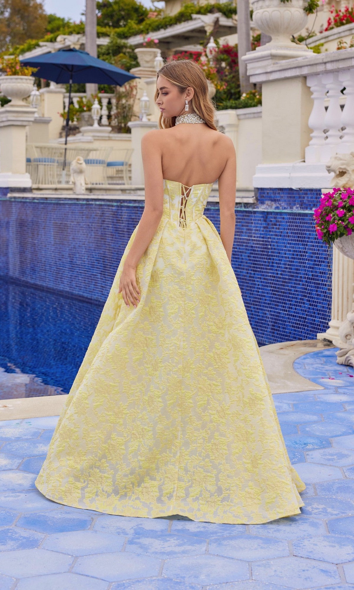 A woman in the Juliet JT2693A Long Prom Dress, a strapless yellow jacquard gown with an A-line skirt and lace-up back, stands by a blue-tiled pool amid classical architecture and flowers, facing away.