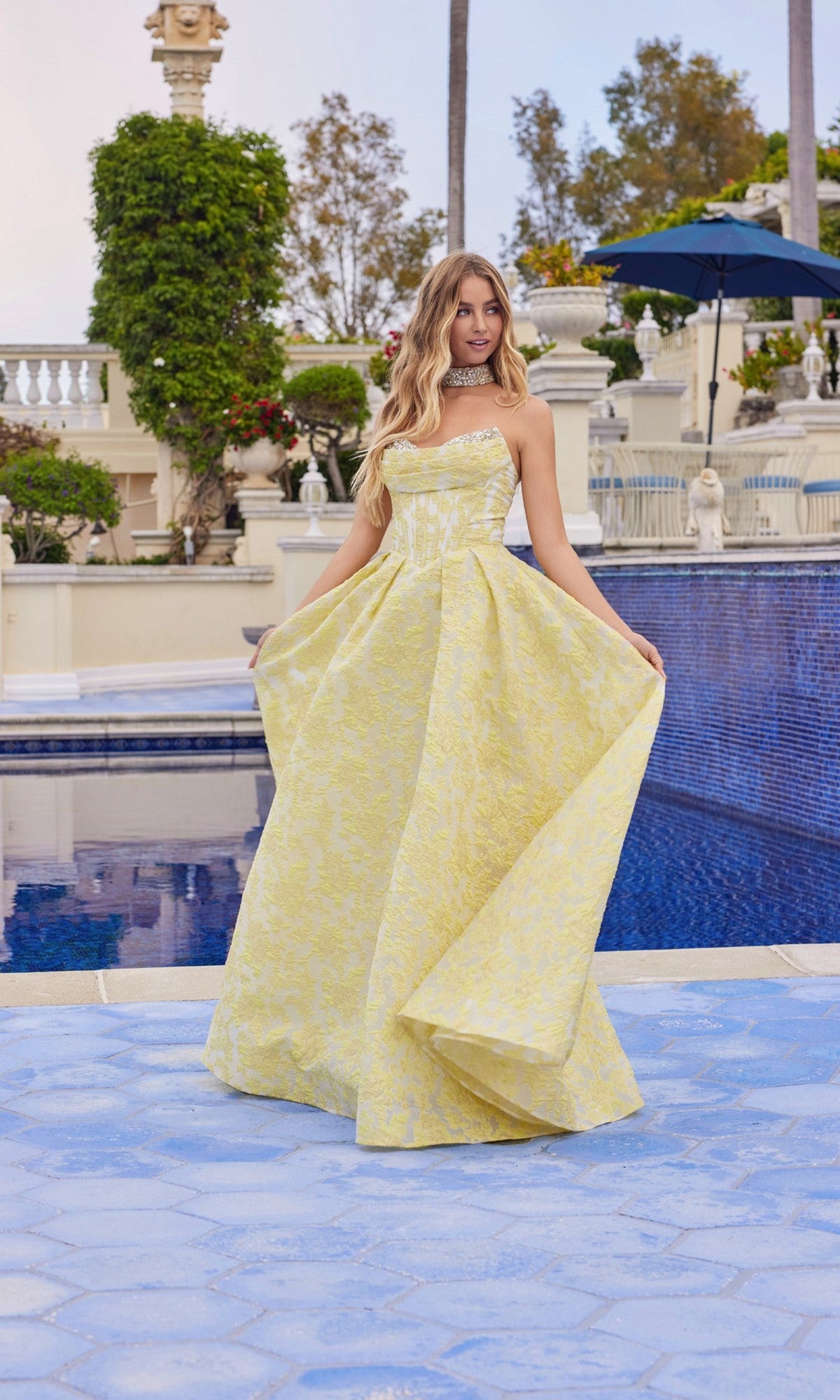 A woman in the yellow Long Prom Dress: Juliet JT2693A with a strapless neckline and a-line skirt stands by a blue-tiled pool, holding out her gown, surrounded by greenery and statues.