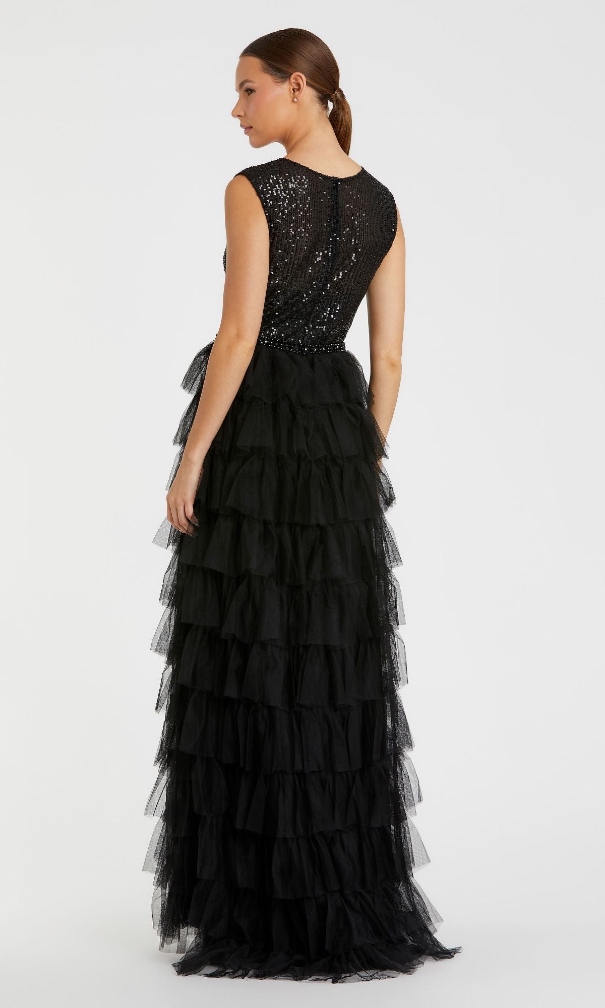 A woman stands facing away in the Mac Duggal 27055 Long Black Ruffled Formal Dress, featuring a sleeveless sequined top and a tiered tulle skirt.