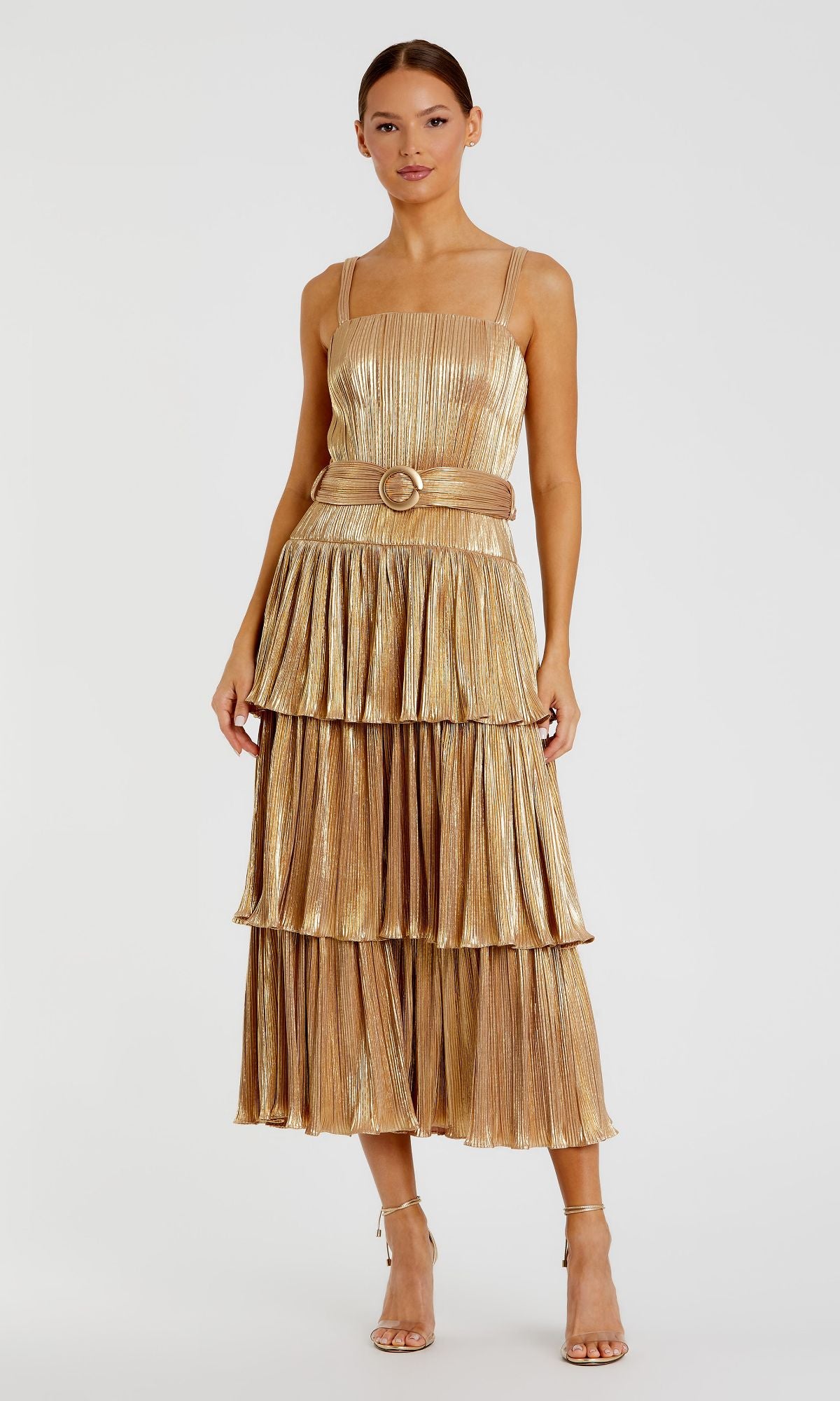 A woman in a Mac Duggal Tea-Length Metallic Party Dress 27062—gold, pleated, tiered, with thin straps and a matching belt—pairs it with strappy heels and stands against a plain white background.