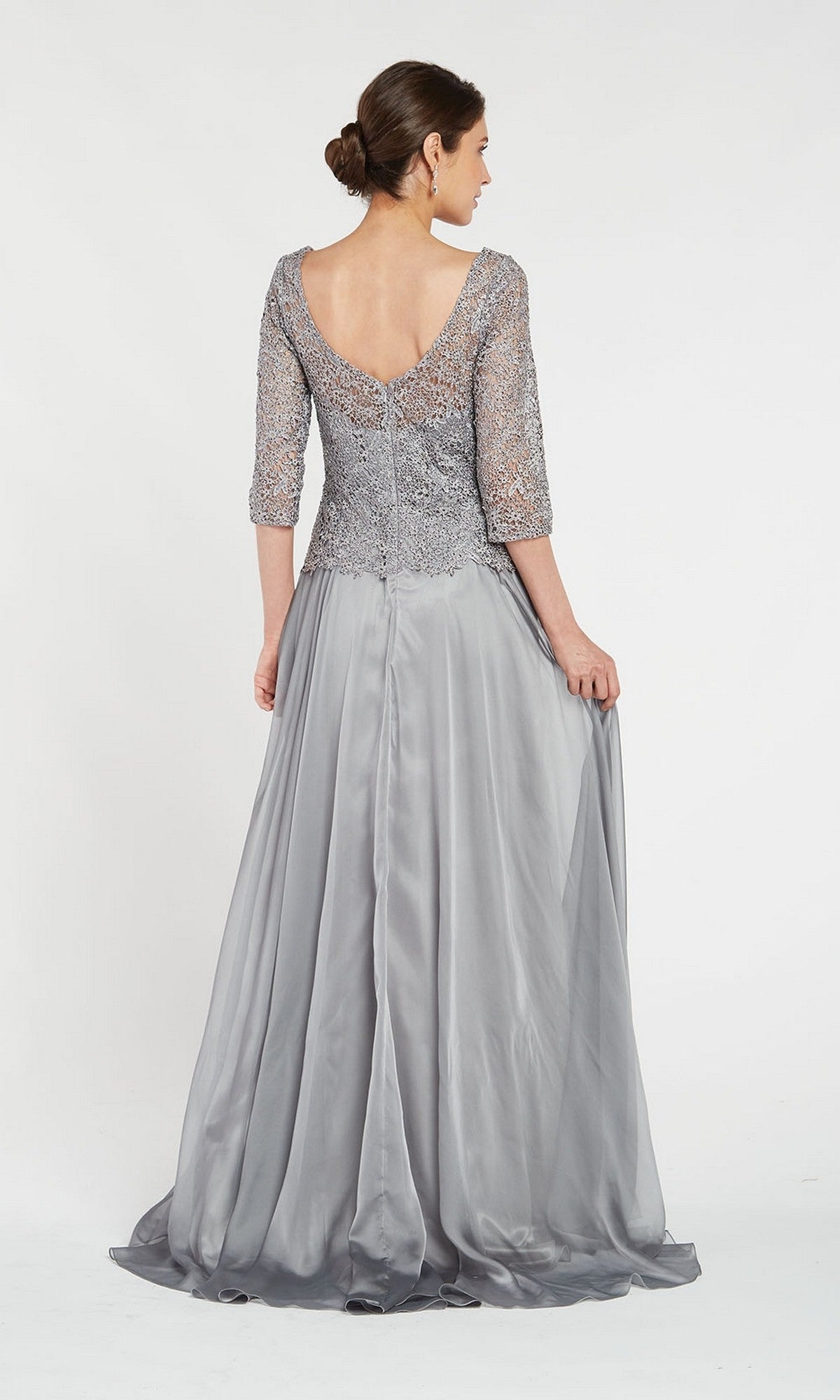 Wearing the Alyce 27251, a mother of the bride stands facing away in a silver-gray long formal dress featuring a lace bodice and sleeves, gently holding her gown’s skirt.