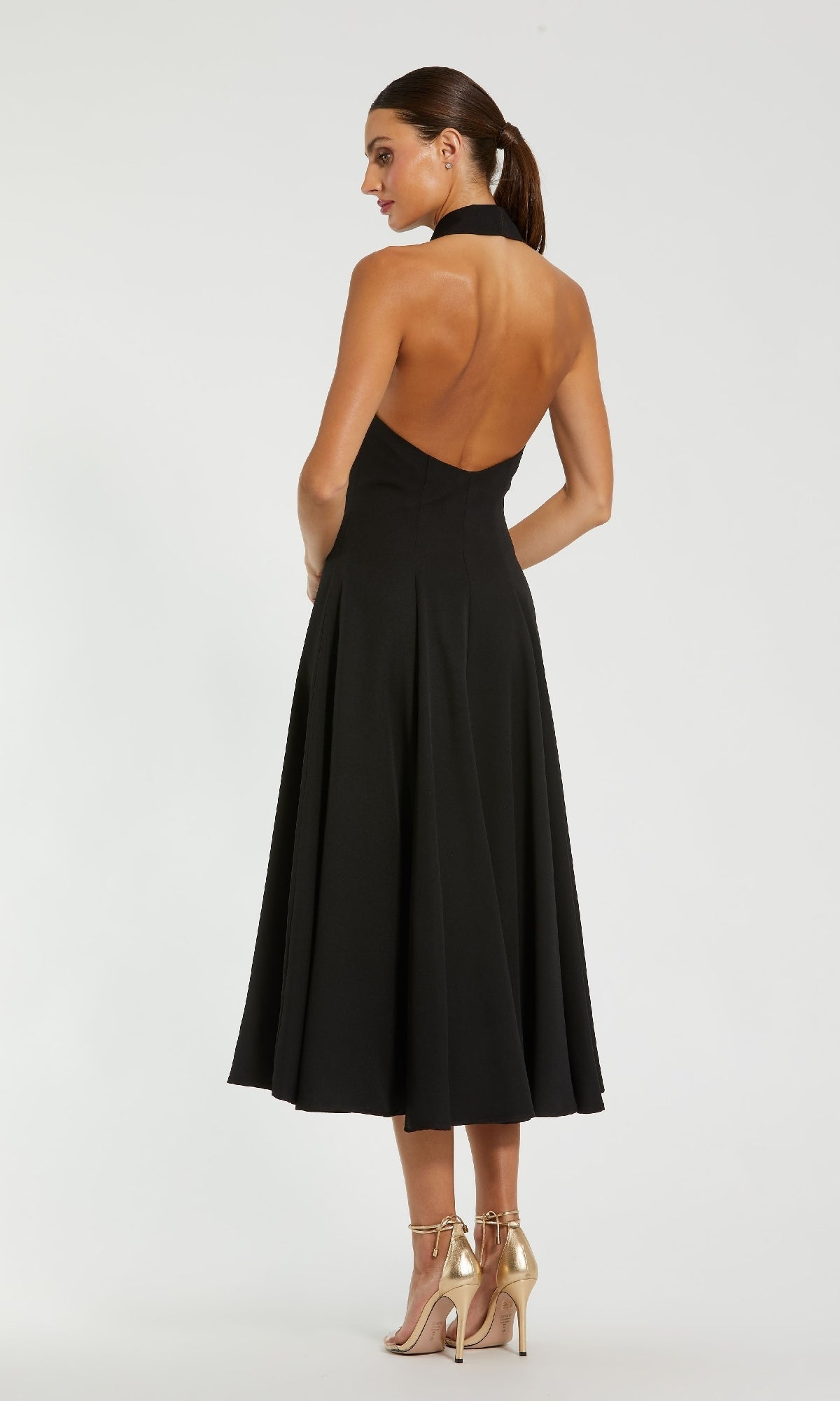 A woman stands against a plain white background wearing the Mac Duggal 27252 Blazer Halter Midi Party Dress, a black open-back halter-neck midi dress, paired with gold high-heeled sandals.