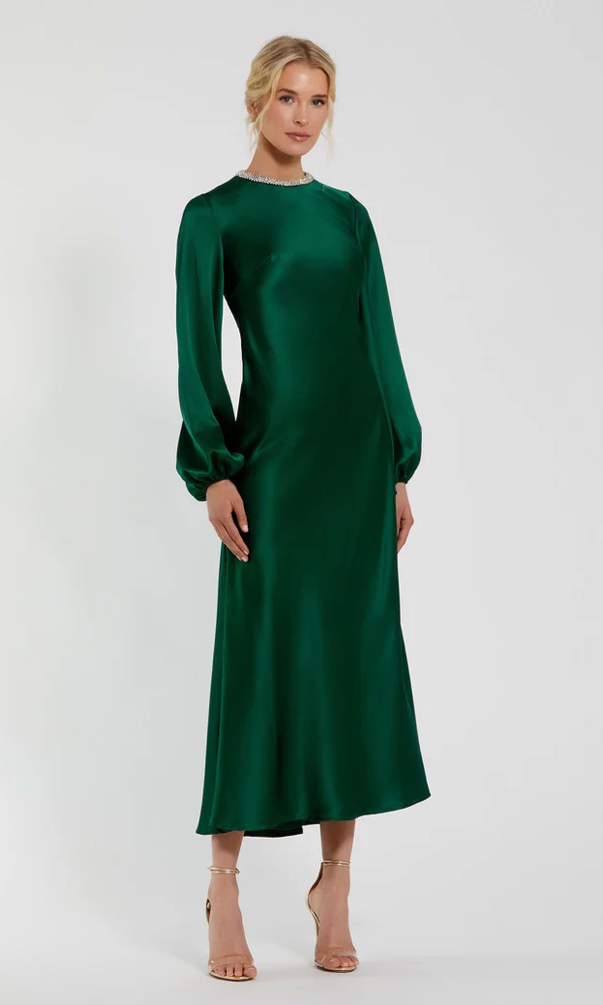 A person stands wearing the Modest Midi Wedding Guest Dress: Mac Duggal 27350, a long-sleeve emerald green satin dress with a beaded high neckline, paired with tan heeled sandals against a plain light background.