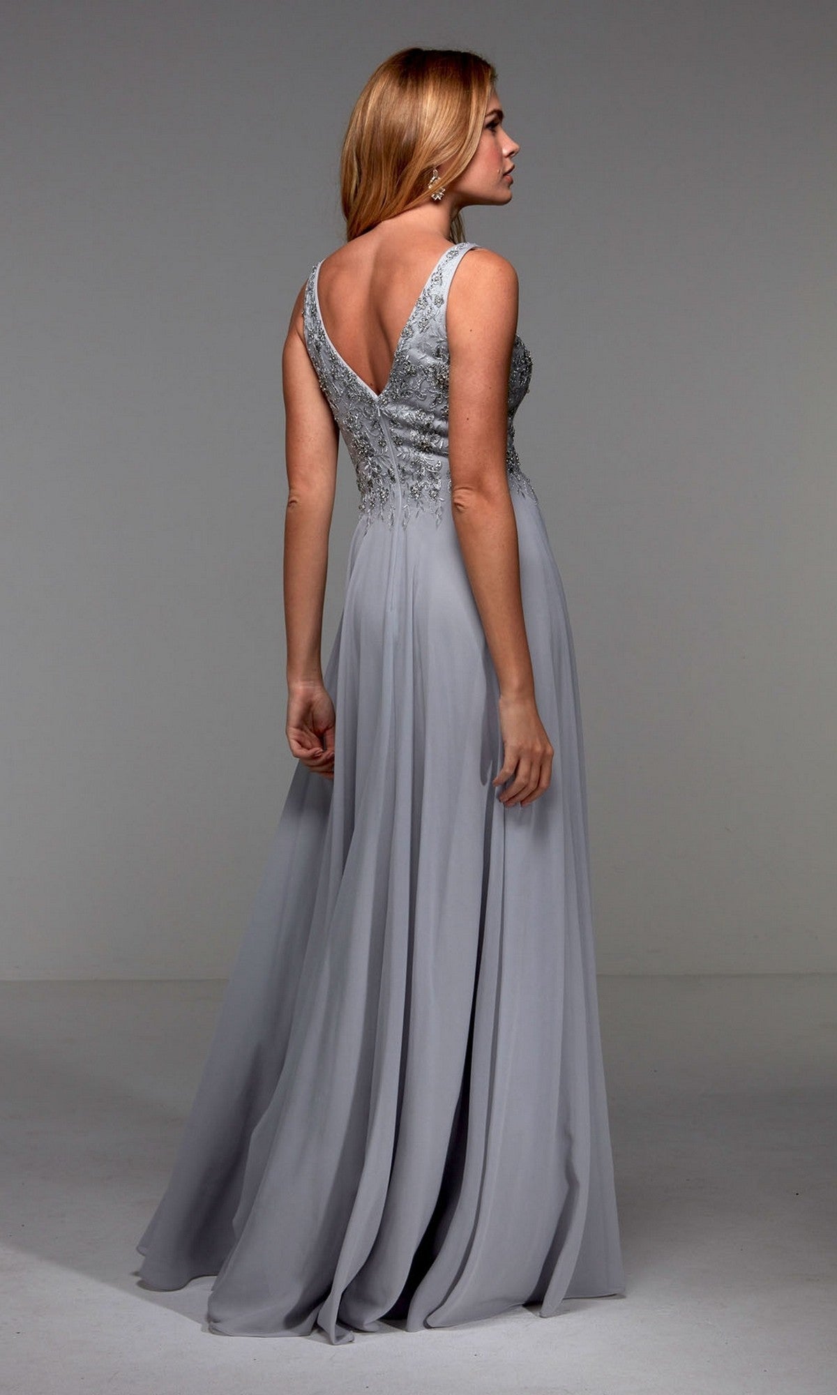 Woman models the Alyce 27473, a sleeveless, floor-length silver chiffon A-line dress with a deep V-back and flowing skirt, standing with her back to the camera.