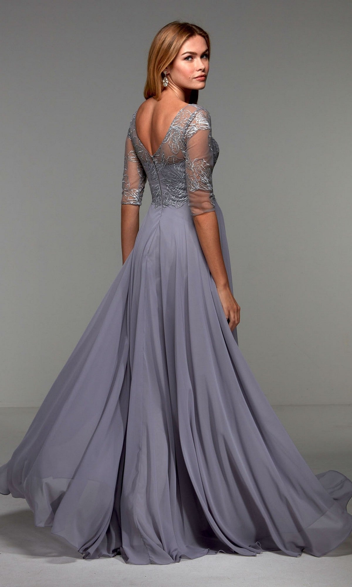 Woman wearing the Alyce 27475 long formal dress—light lavender, floor-length, with sheer lace sleeves and a low back—stands looking over her shoulder. An elegant option for formal events or as a mother-of-the-bride dress.