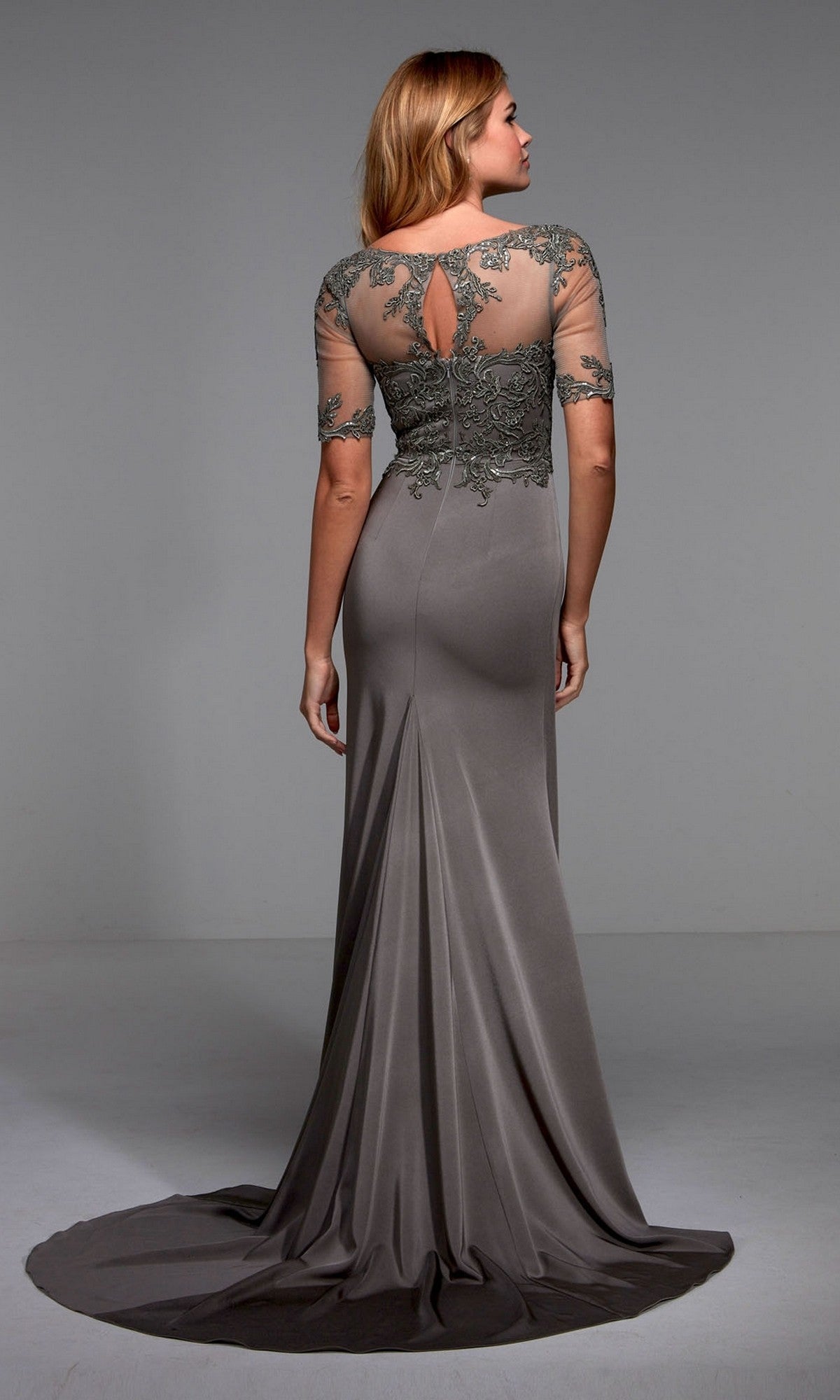 A woman stands facing away from the camera in a plain studio, wearing the Alyce 27477 long formal dress—floor-length, taupe, with an embellished bodice and sheer, embroidered back and sleeves.