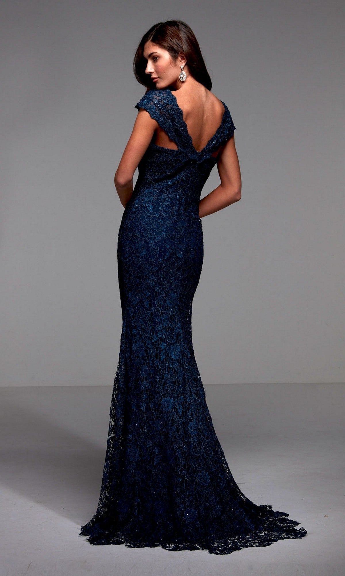 A woman wears the Alyce 27496 long formal dress—a dark blue, floor-length mermaid gown made of shimmering lace with a low back and cap sleeves—standing against a plain background.