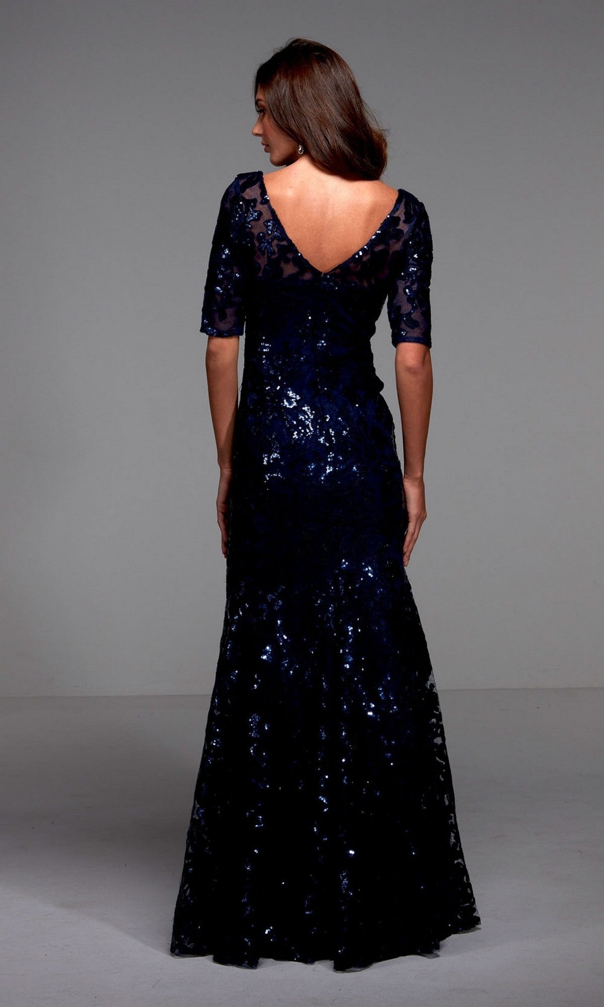 Model in Alyce 27497—a dark blue, floor-length formal dress with shimmering sequins, half sleeves, and a V-back—stands against a plain background. An elegant choice ideal for a mother-of-the-bride.