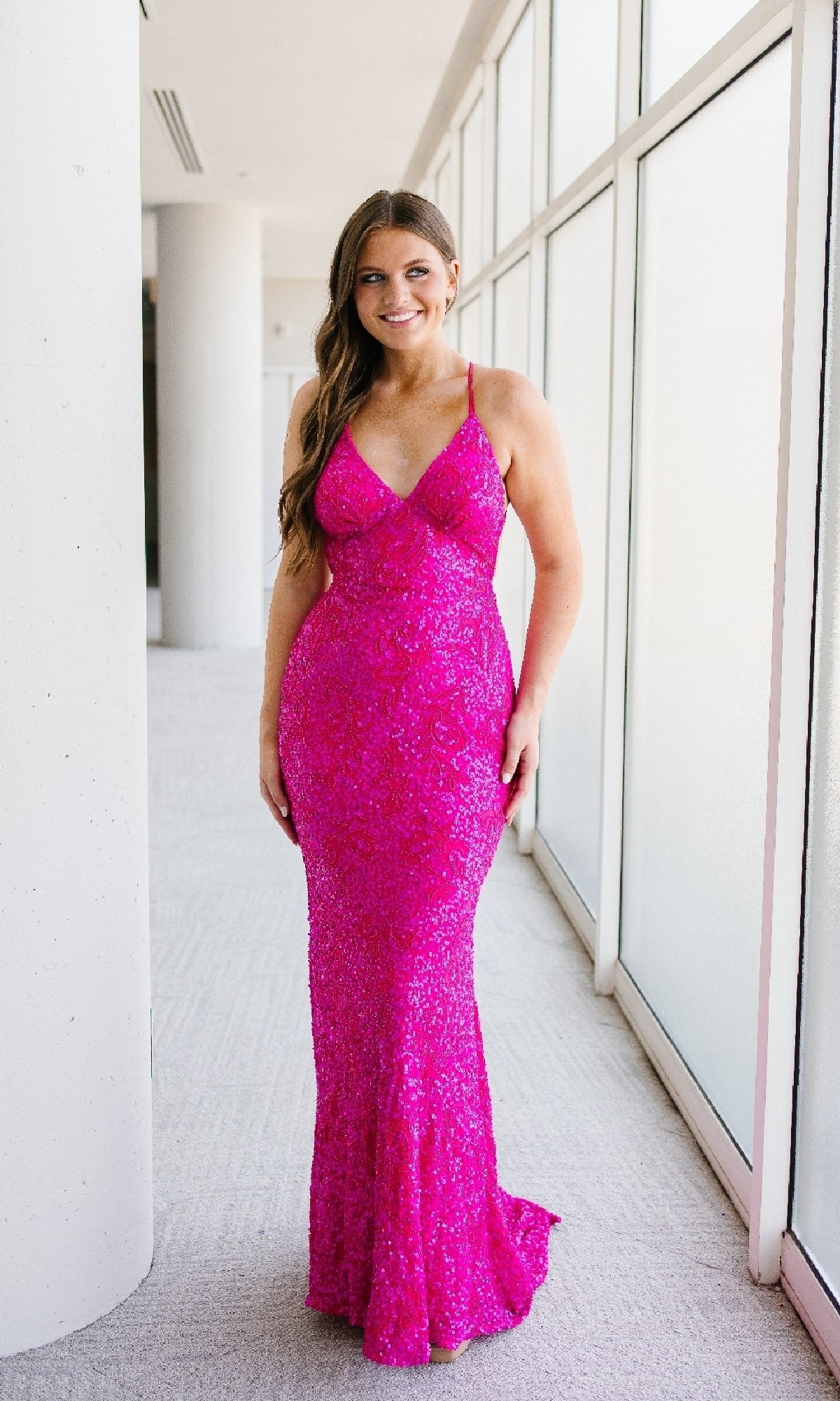 A woman stands indoors by large windows, wearing the Sleek Fitted Long Sequin Prom Dress by Aleta—a floor-length, bright pink gown with thin straps and shimmering sequins.