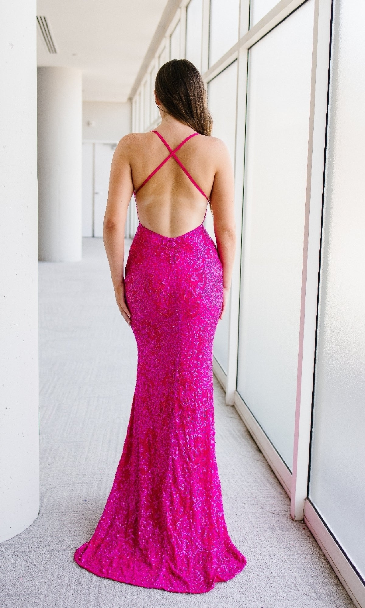A woman stands indoors facing away, wearing the Sleek Fitted Long Sequin Prom Dress by Aleta— a floor-length, bright pink gown featuring crisscross straps and a stunning backless design.
