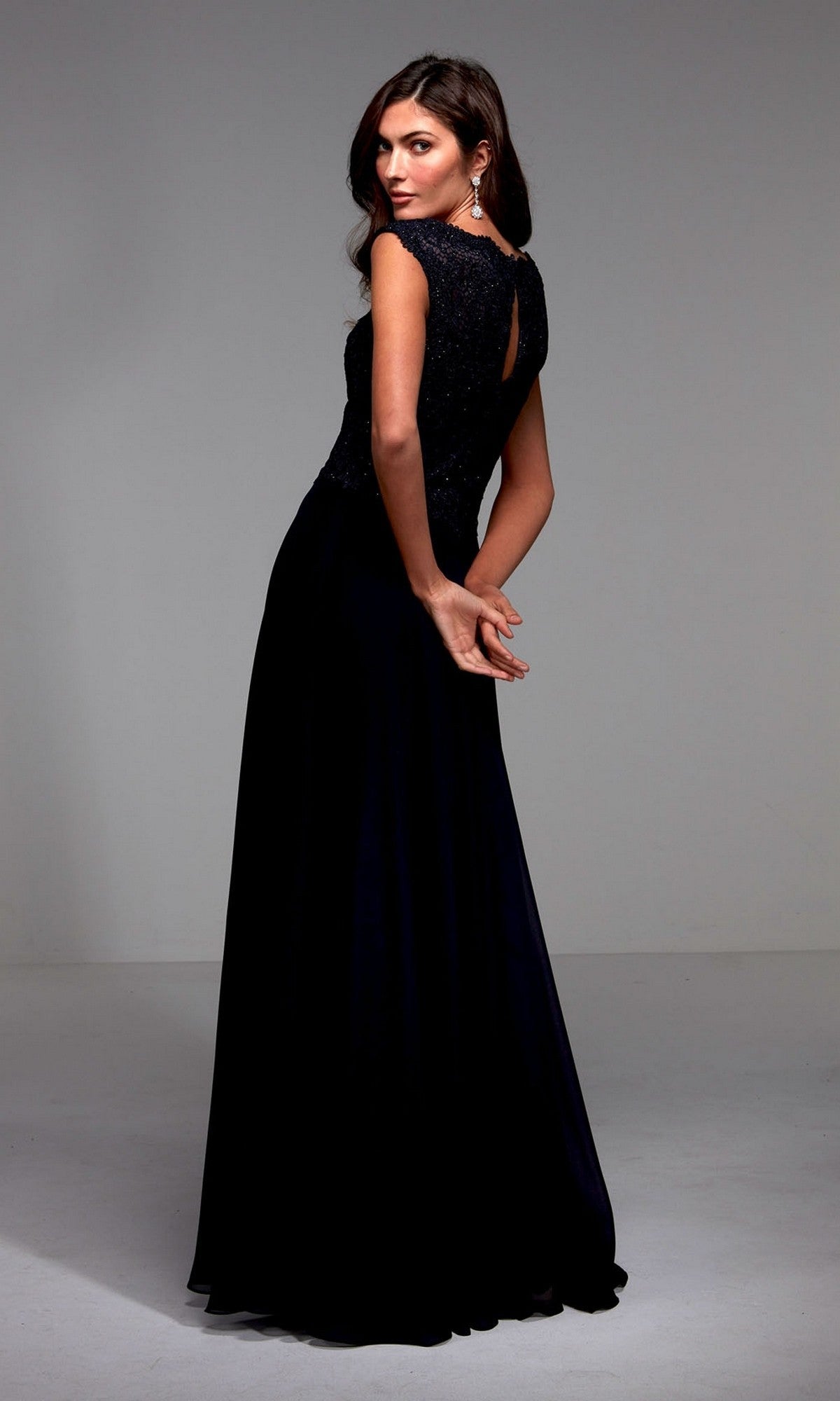 A woman wearing the Alyce 27504 long formal dress—an elegant A-line chiffon gown with a lace bodice—stands with her back partially turned and hands clasped behind her in a plain studio setting.